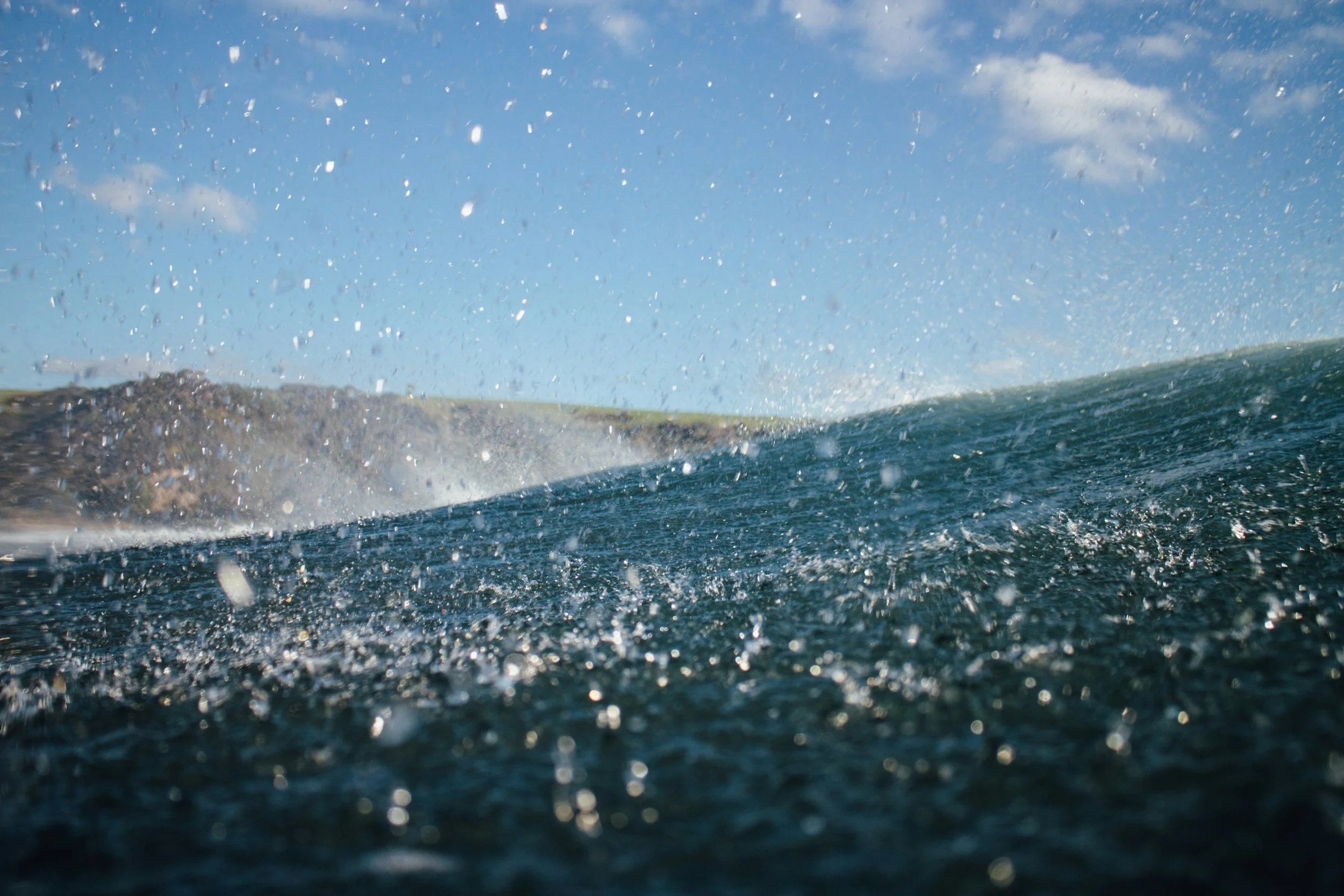 Close-up of ocean waves with water droplets flying in the air under a clear blue sky, with a distant landmass in the background.