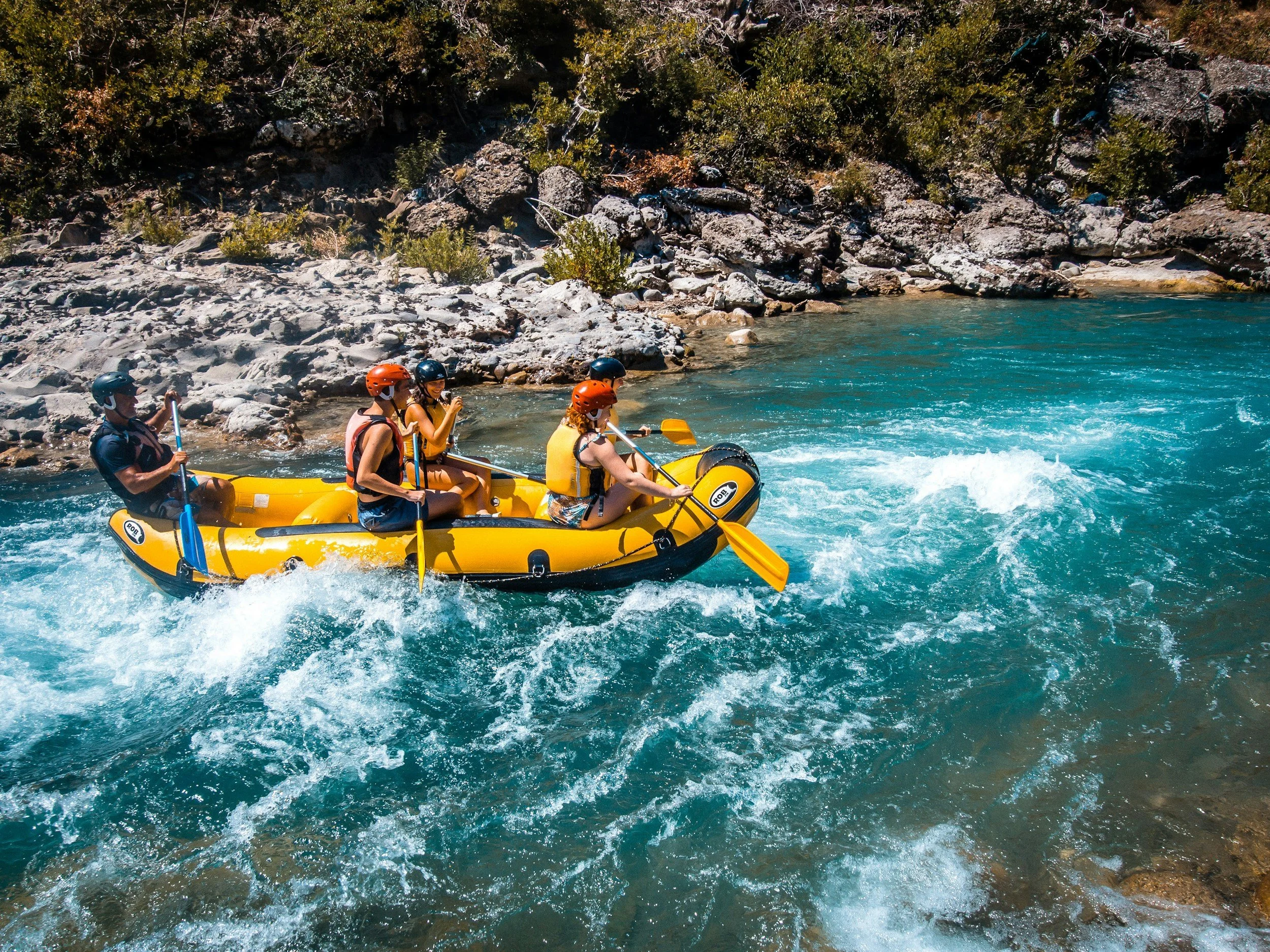 Four people wearing helmets and life jackets are paddling a yellow inflatable raft on a river with rocky banks and flowing water.
