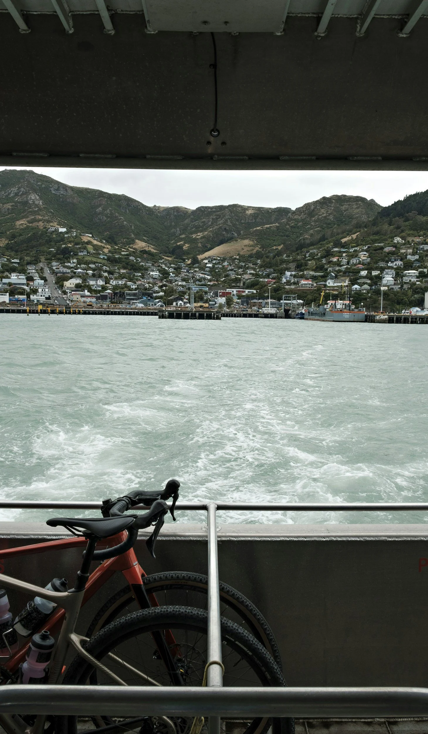 View from the back of a boat showing a mountain town and harbor, with two bicycles at the bottom of the image and a building overhead.