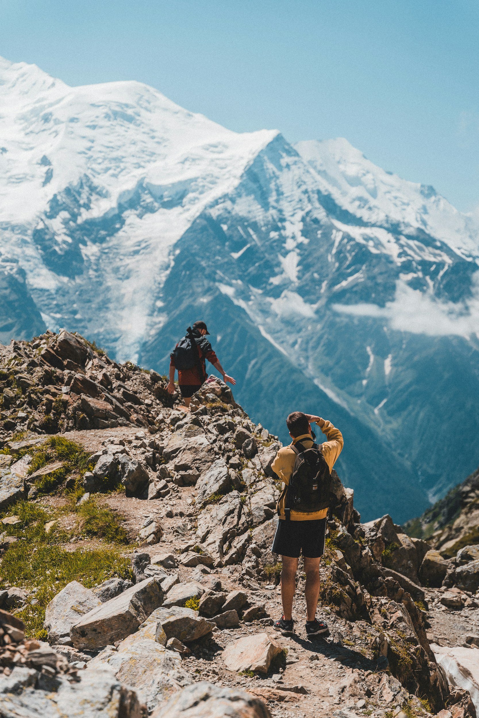 Three hikers are trekking along a rocky mountain trail in front of a tall snow-capped peak under a clear blue sky.
