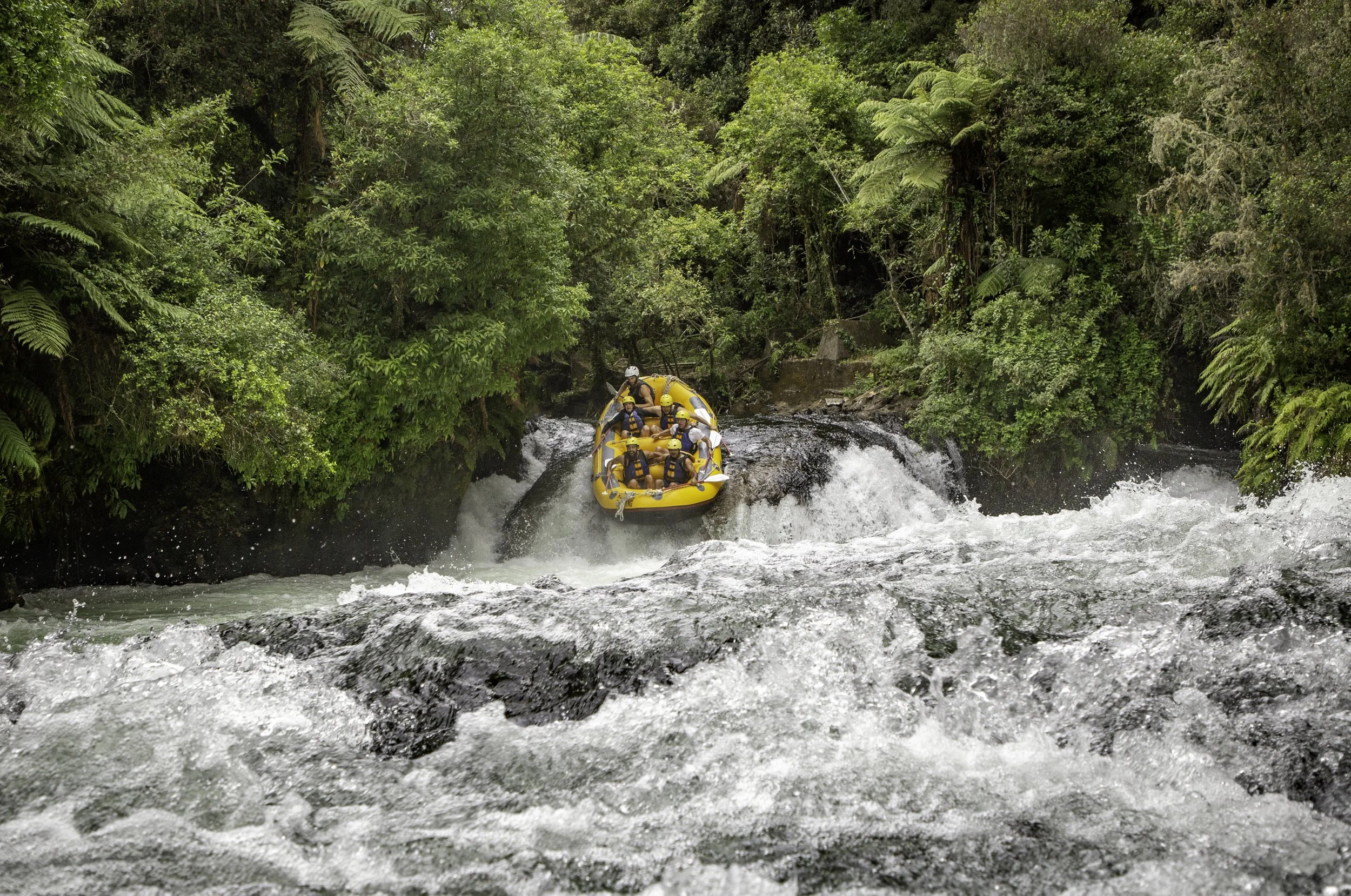 People on a yellow raft white water rafting on a river surrounded by lush green trees and foliage.
