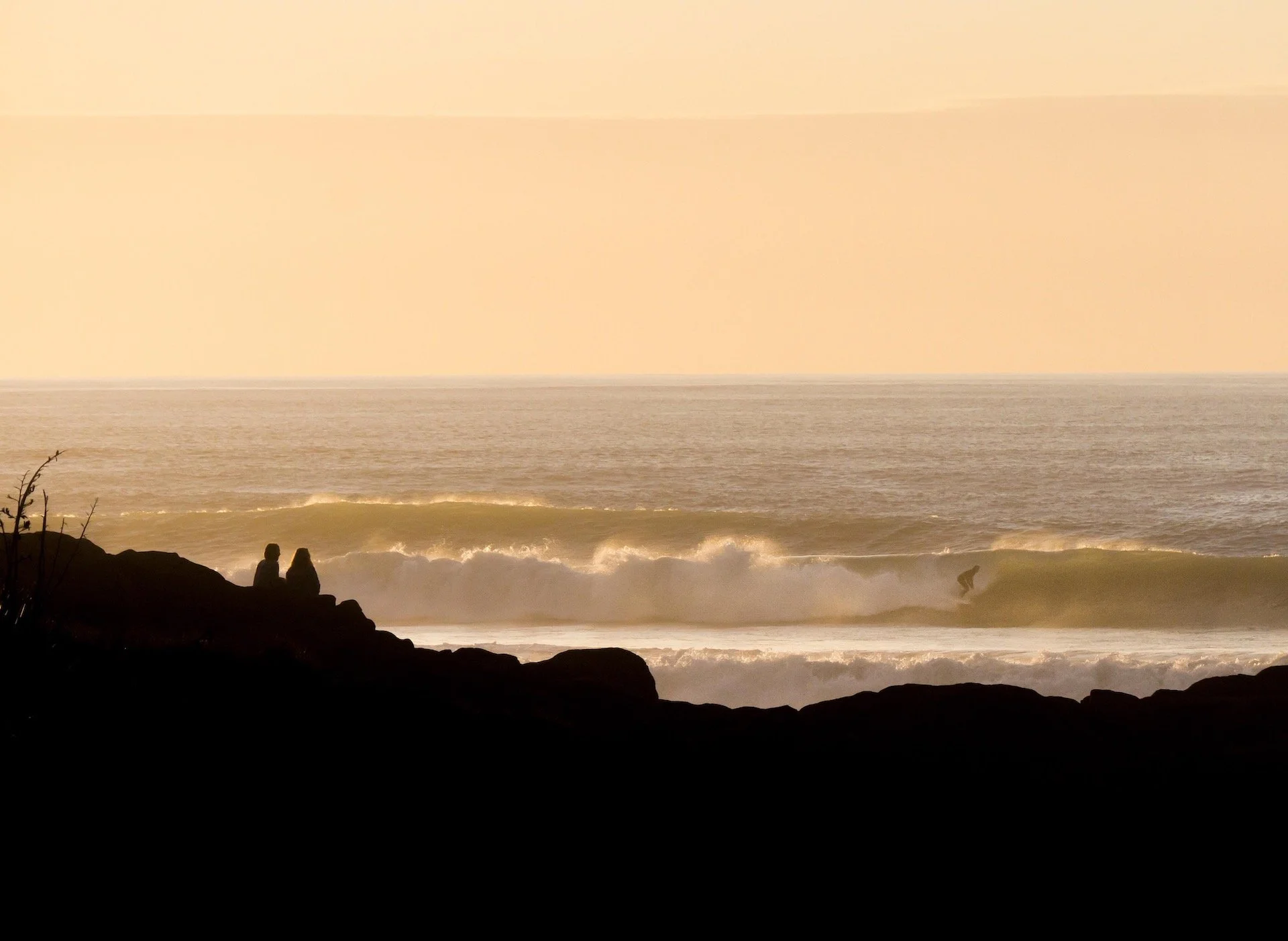 Silhouettes of two people sitting on rocks watching a surfer riding a wave at sunset.