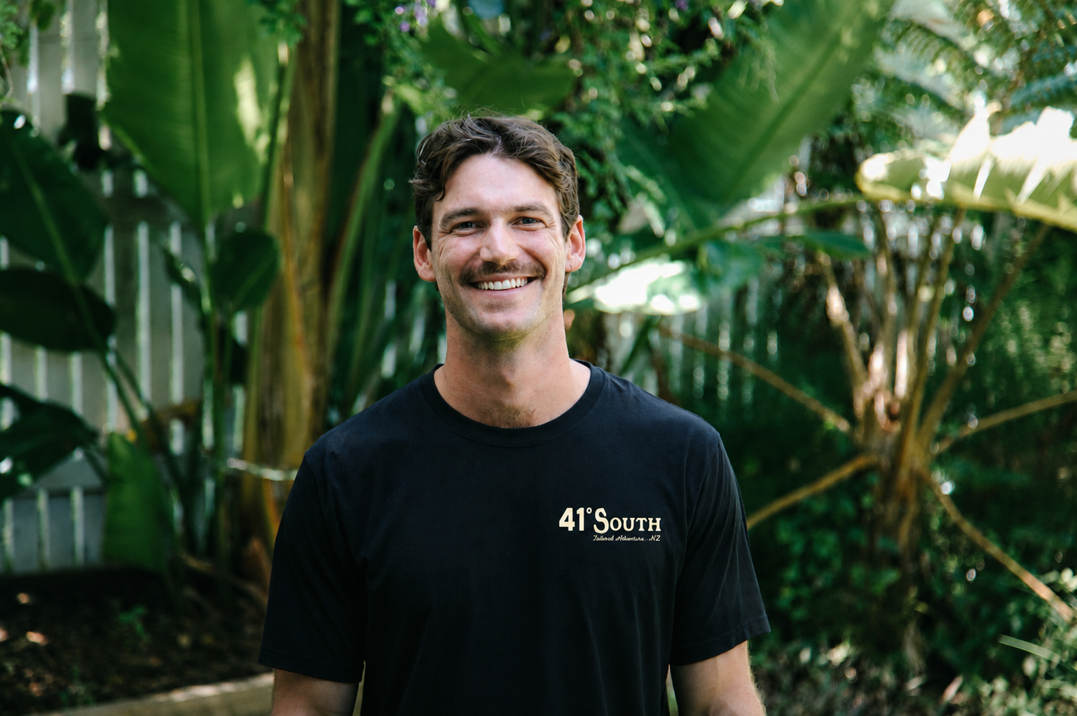 A smiling man with dark wavy hair and a mustache, wearing a black T-shirt, standing outdoors with lush green tropical plants in the background.