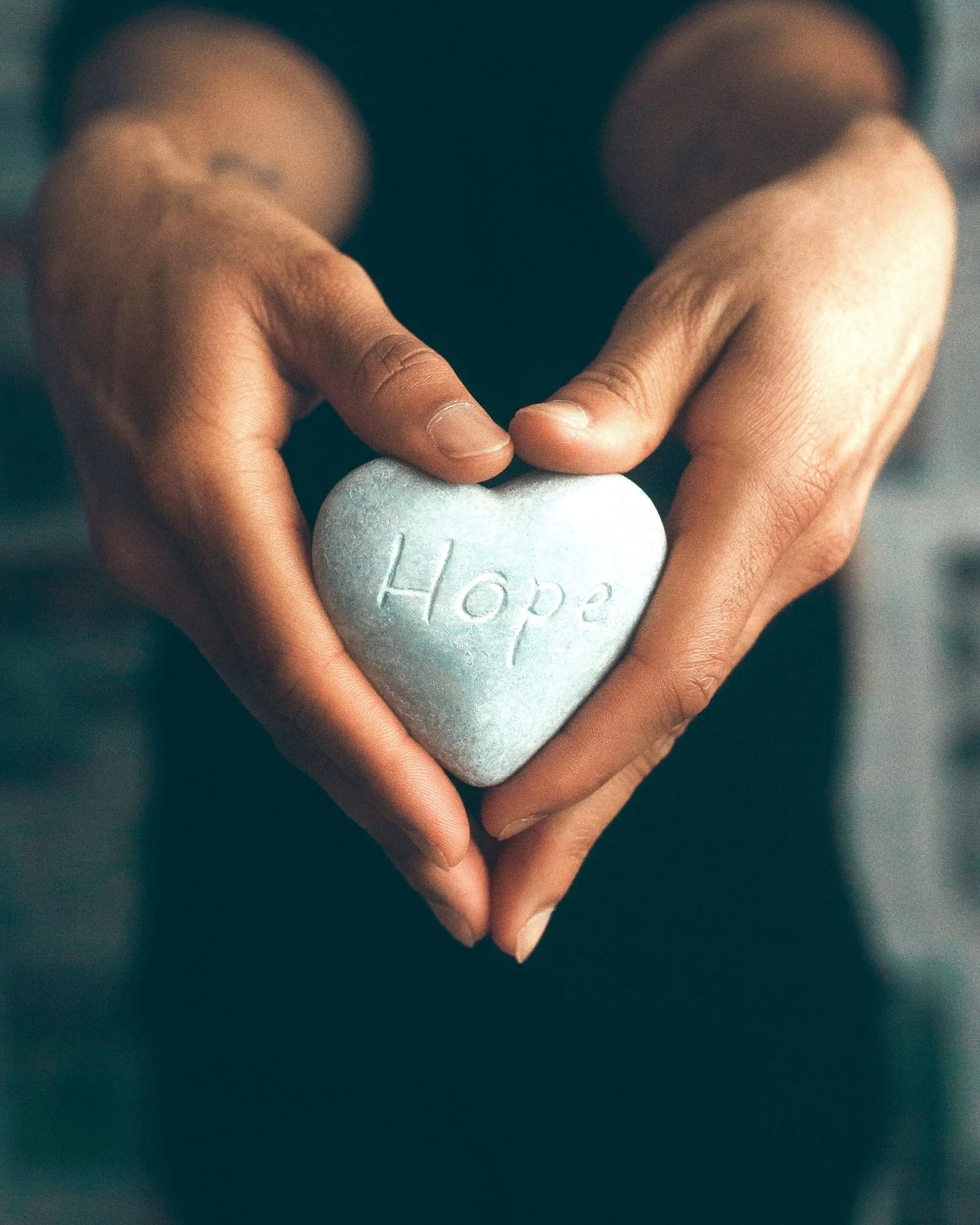 Hands forming a heart shape around a heart-shaped stone engraved with the word 'Hope'.
