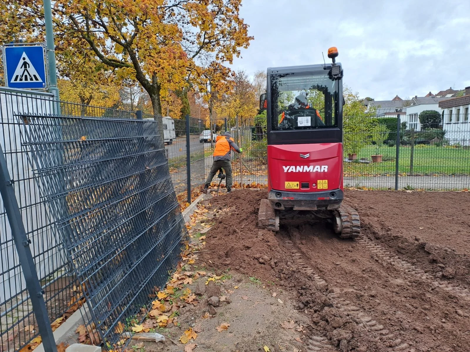 Ein Baulkwerk mit einem roten Yanmar Minibagger beim Graben einer Baustelle, während ein Arbeiter in orangefarbener Weste im Hintergrund die Arbeiten durchführt. Es gibt eine Metallzaun, Bäume mit Herbstlaub und Häuser im Hintergrund.