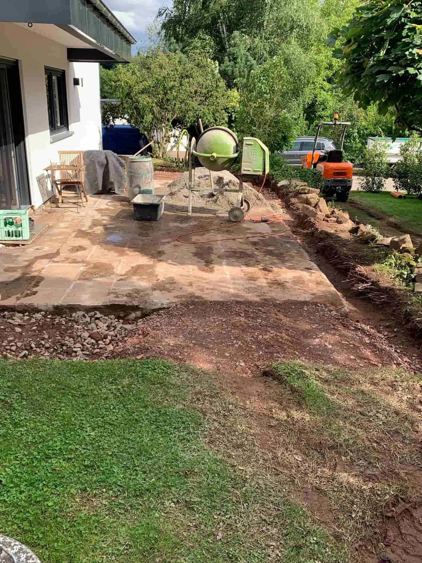Eine Terrasse, die gerade umgestaltet wird, mit Baustellengeräten wie Betonmischer, Schaufel und Bauwerkzeugen. Der Bereich ist teilweise aufgerissen, um den Boden zu erneuern. Im Hintergrund sind Bäume und ein Fahrzeug zu sehen.