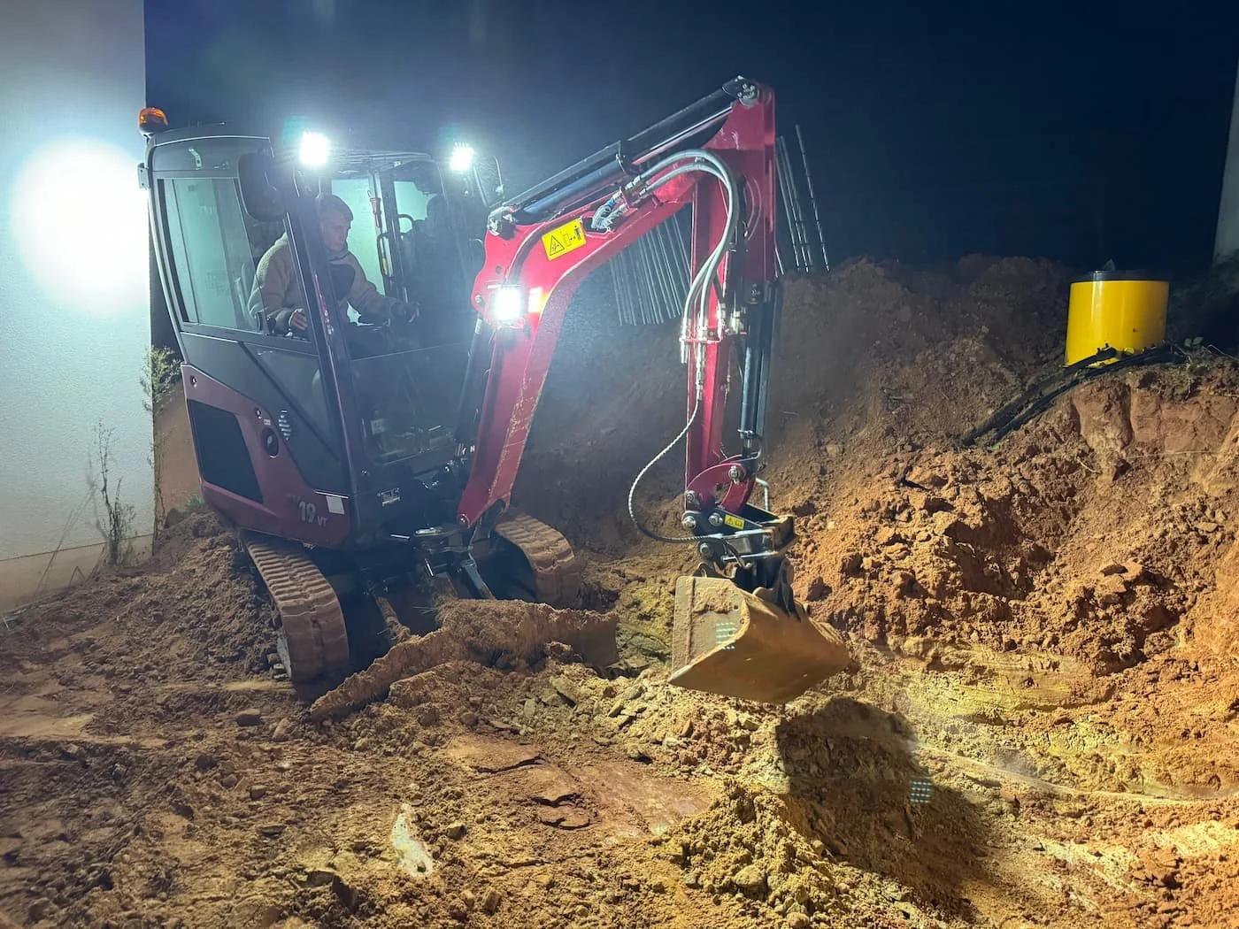 Ein Minibagger hebt Erde bei Nacht, mit Scheinwerfern beleuchtet, auf einer Baustelle.
