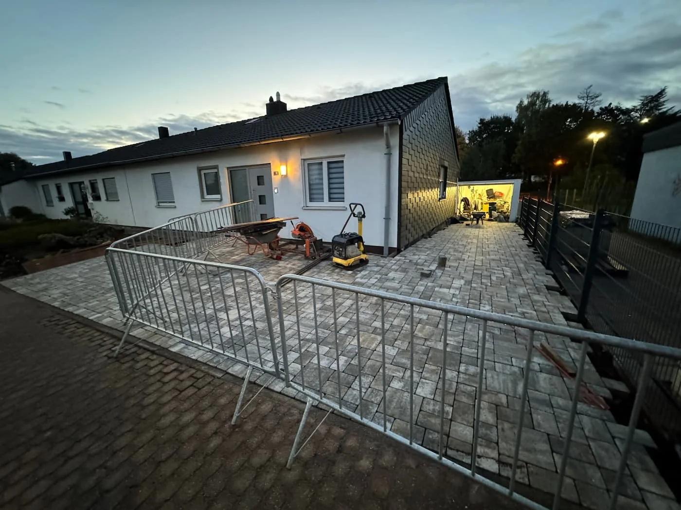 Ein Haus bei Dämmerung, im Vorgarten werden Pflasterarbeiten durchgeführt. Es gibt Baufahrzeuge, Werkzeug und Bauzäune, die den Arbeitsbereich absperren.
