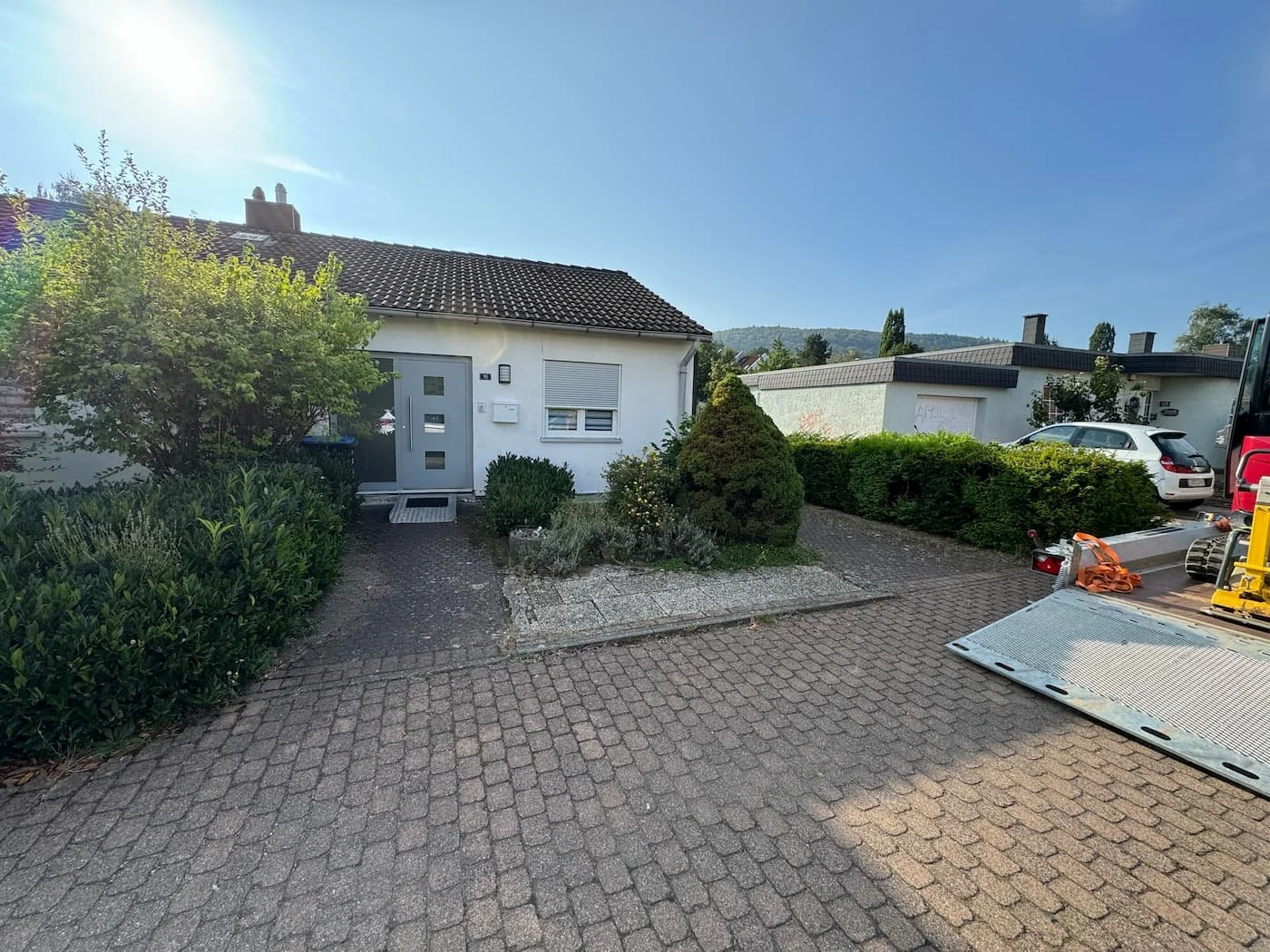 Vorderansicht eines weißen Einfamilienhauses mit grauer Tür und Garten, umgeben von Büschen und kleinen Bäumen, mit einem gepflasterten Vorplatz, einem Van und einem abgestellten LKW auf dem Hof bei sonnigem Himmel.