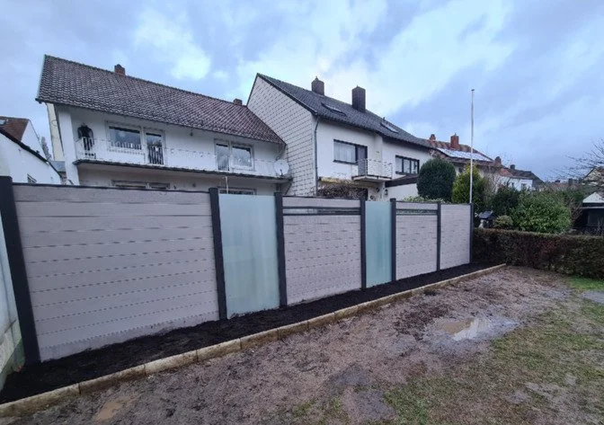 Ein neuer, gemauerter Lattenzaun zwischen Häusern, mit einer Rasen- und Flächen aus Erde davor, bei bewölktem Himmel.