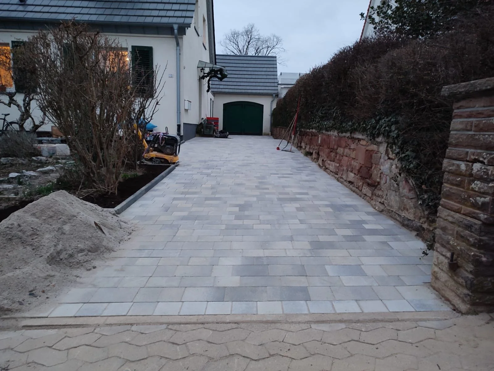 Ein fertiggestellter gepflasterter Weg vor einem Haus mit Garten auf der linken Seite und einer Hecke auf der rechten Seite, im Hintergrund ein Garagentor, der Himmel ist bewölkt.