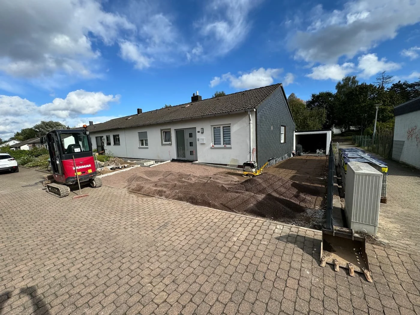 Bauarbeiten vor einem Wohnhaus mit Baugerät und Erde, die für eine neue Terrasse oder Einfahrt vorbereitet wird.