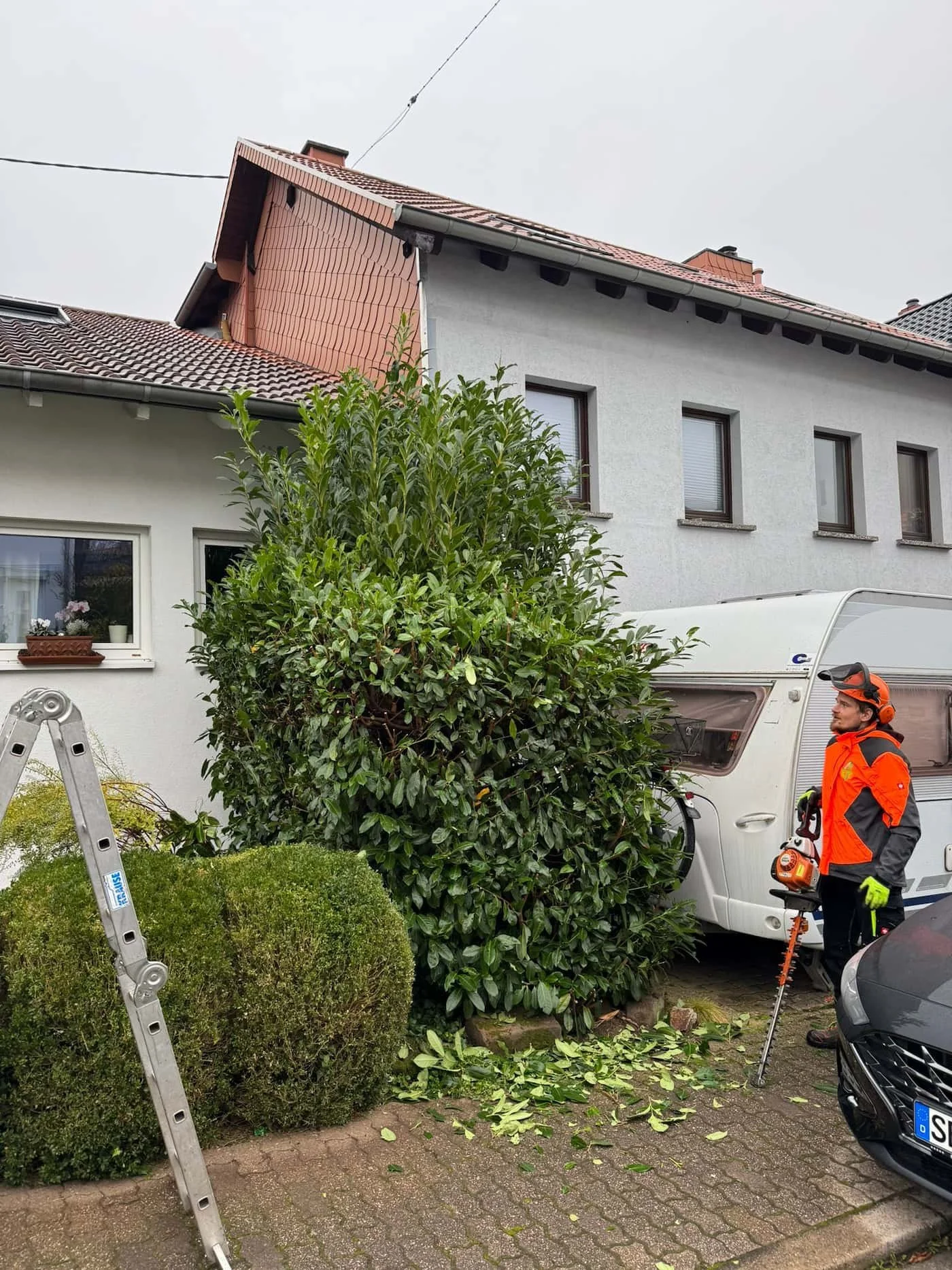 Ein Gärtner steht neben einem umliegenden, umgestürzten Busch vor einem Wohnhaus, während er das herabgestürzte Laub auf einem gepflasterten Hof mit einem Kettenrasenmäher sammelt.