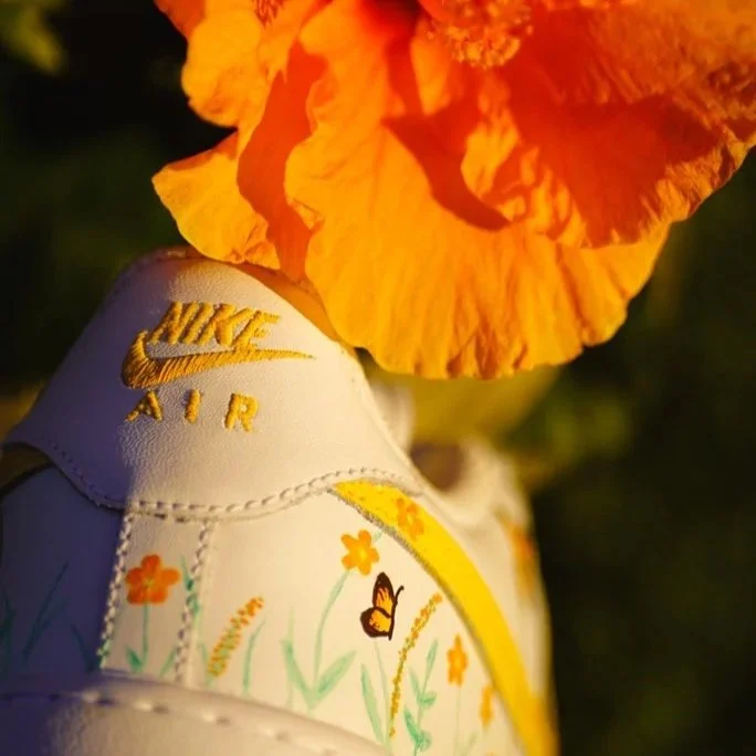 A white Nike Air sneaker decorated with orange flowers and a butterfly, with an orange hibiscus flower partially covering the top of the shoe.