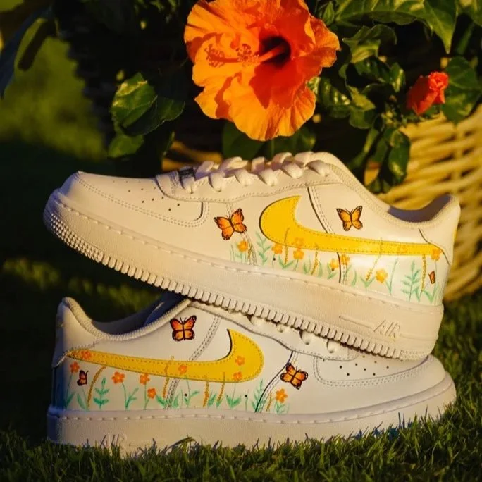Pair of white Nike Air Force 1 sneakers with orange hand-painted floral and butterfly designs, displayed outdoors near blooming orange hibiscus.