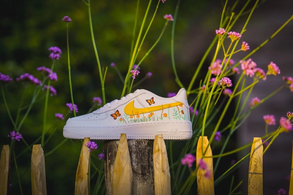 A white Nike Air Force 1 sneaker decorated with orange and yellow butterflies and floral designs, placed on a wooden fence amid flowers.