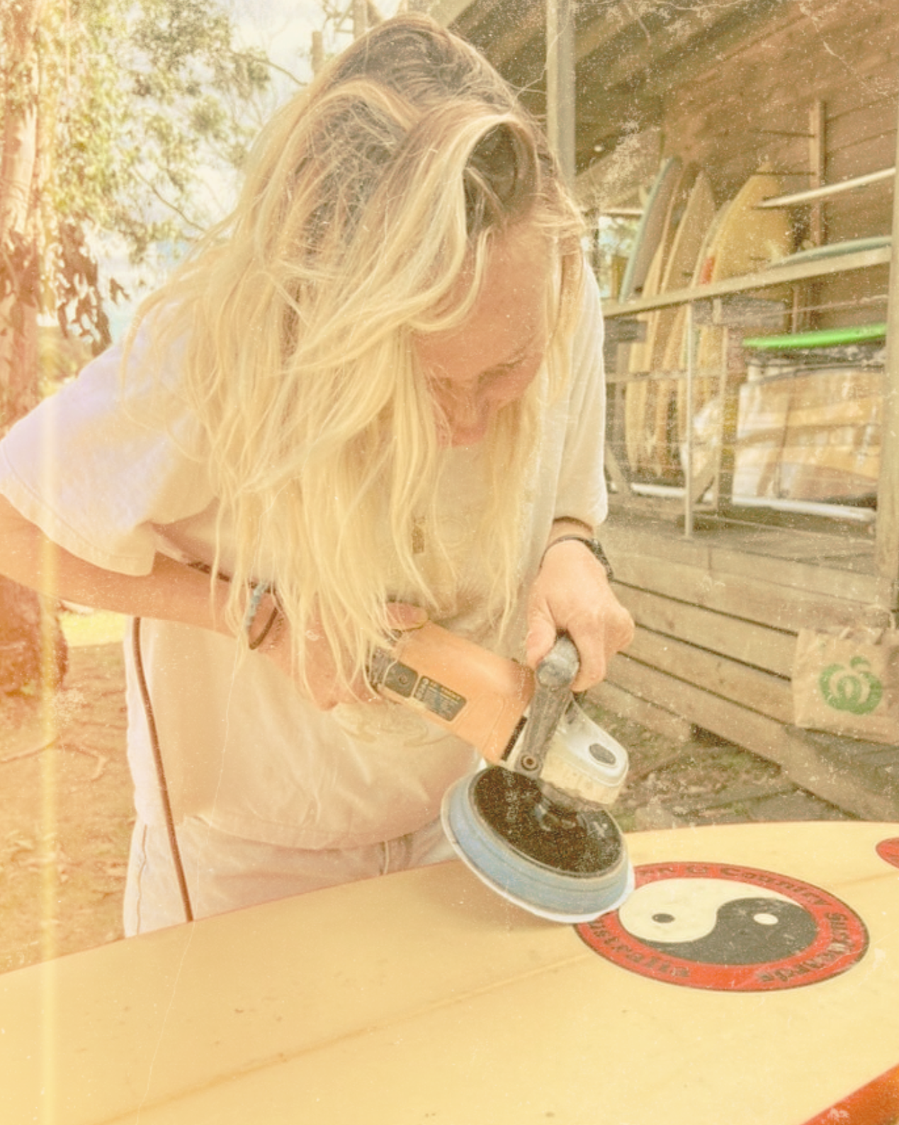 Person fixing a ding in a surfboard, a surfboard, in a workshop or outdoor setting.