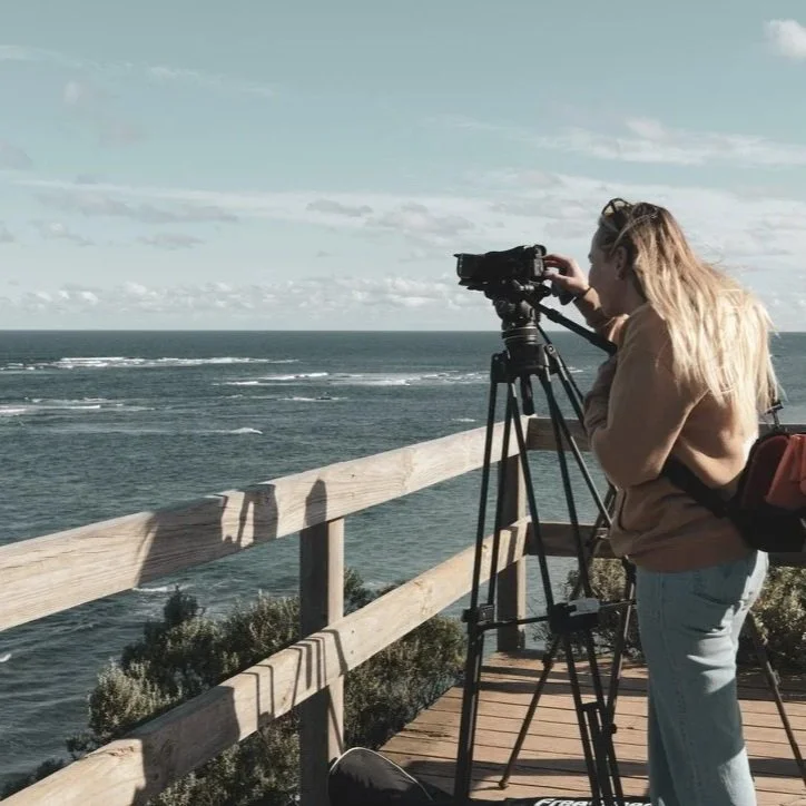Rox, a woman with long blonde hair, is operating a professional video camera on a tripod on an outdoor wooden observation deck overlooking the ocean.