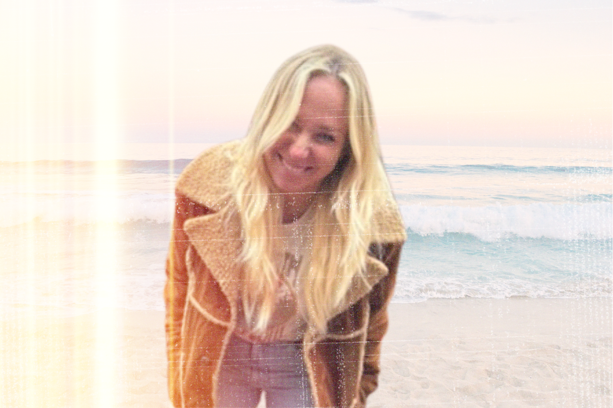 A woman, Rox,  with long blonde hair, smiling at the camera on a beach with waves, at sunset or sunrise.