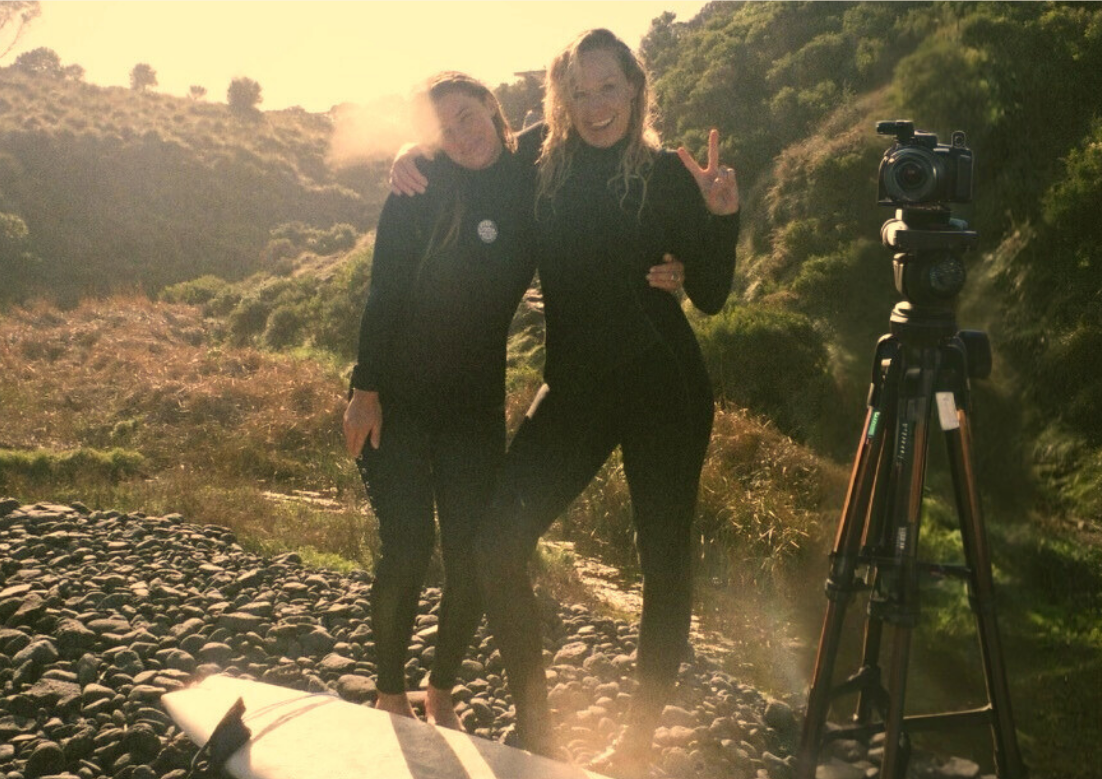Two women smiling and posing in wetsuits at the beach near a camera on a tripod with a rocky trail and green hills in the background. One woman Rox, is making a peace sign.
