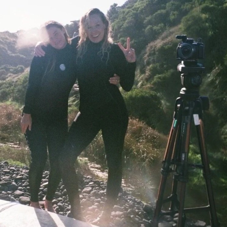 Two women smiling and posing in wetsuits at the beach near a camera on a tripod with a rocky trail and green hills in the background. One woman Rox, is making a peace sign.