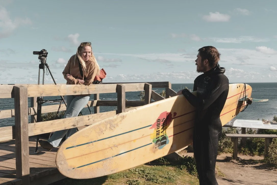 A woman, Rox, is discussing the surf coaching session with a man holding a surfboard on a wooden deck near the The man has short hair and is wearing a black swimsuit. There is a camera on a tripod in the background, and the ocean and sky are visible.