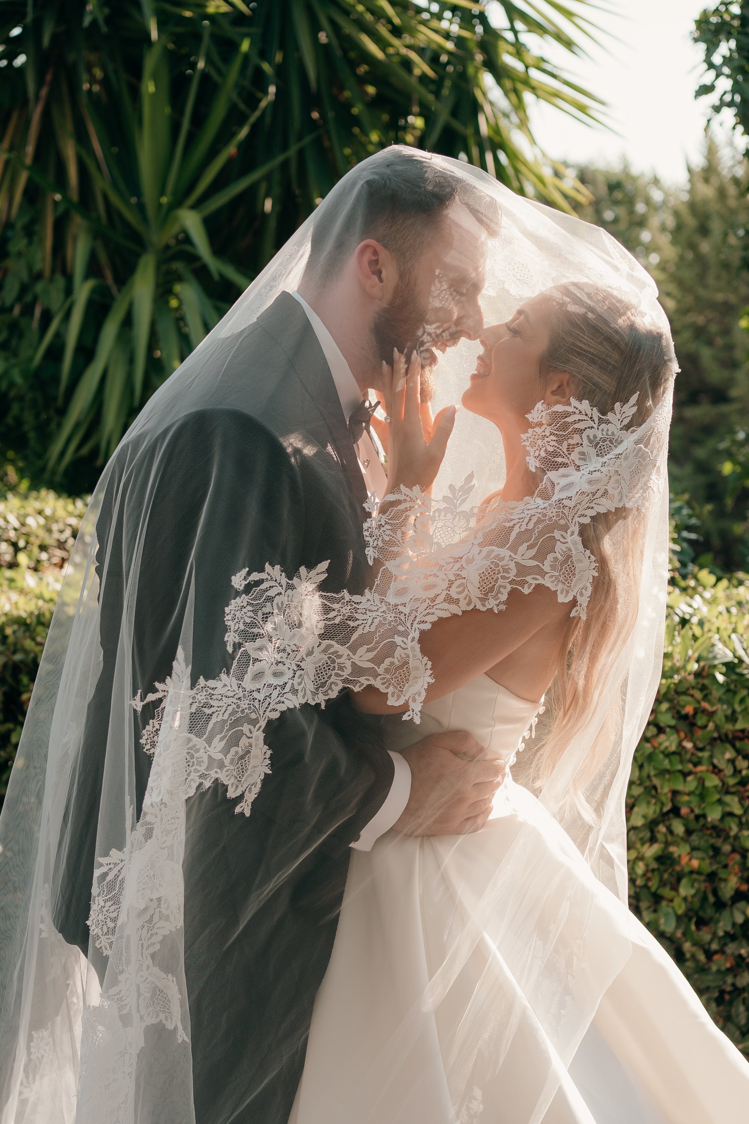 A bride and groom closely embracing under a wedding veil, outdoors with green foliage in the background.