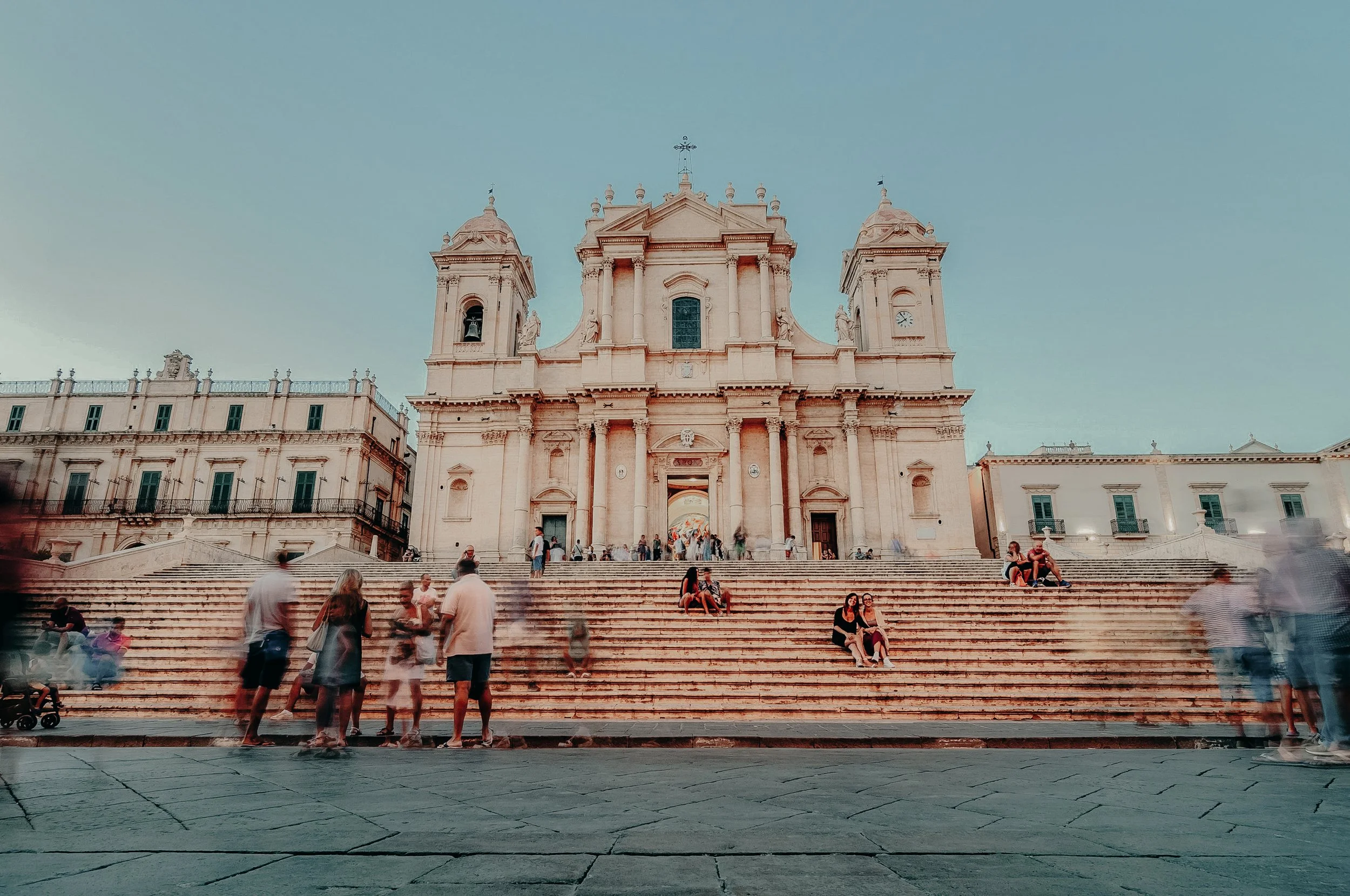 People sitting and walking on the steps in front of a large historic church with twin towers and a central facade, under a clear sky.