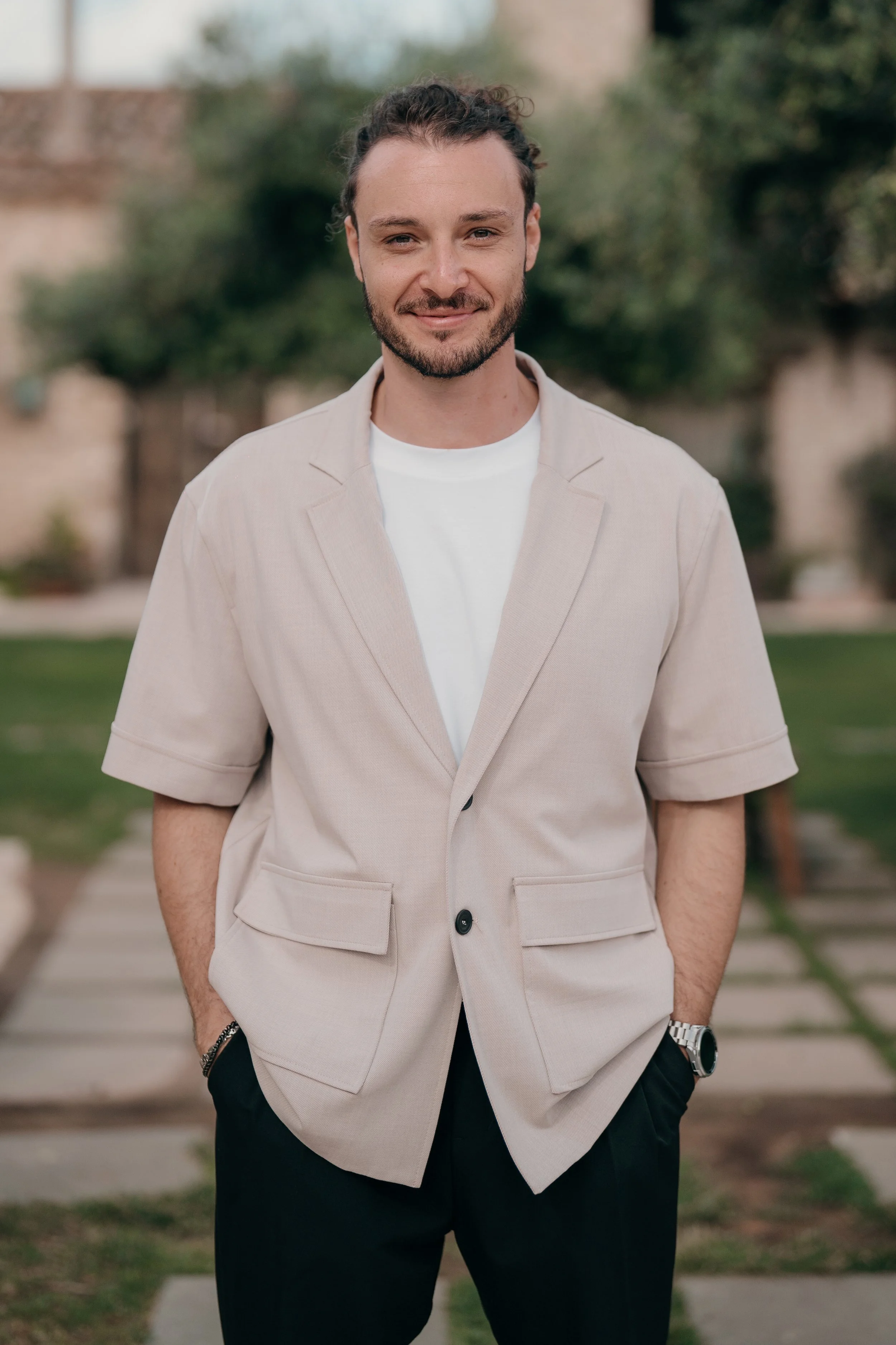 Emanuele Manili standing outdoors on a pathway, smiling, wearing a beige short-sleeve blazer over a white t-shirt, black pants, a wristwatch, and a bracelet, with greenery and houses blurred in the background.