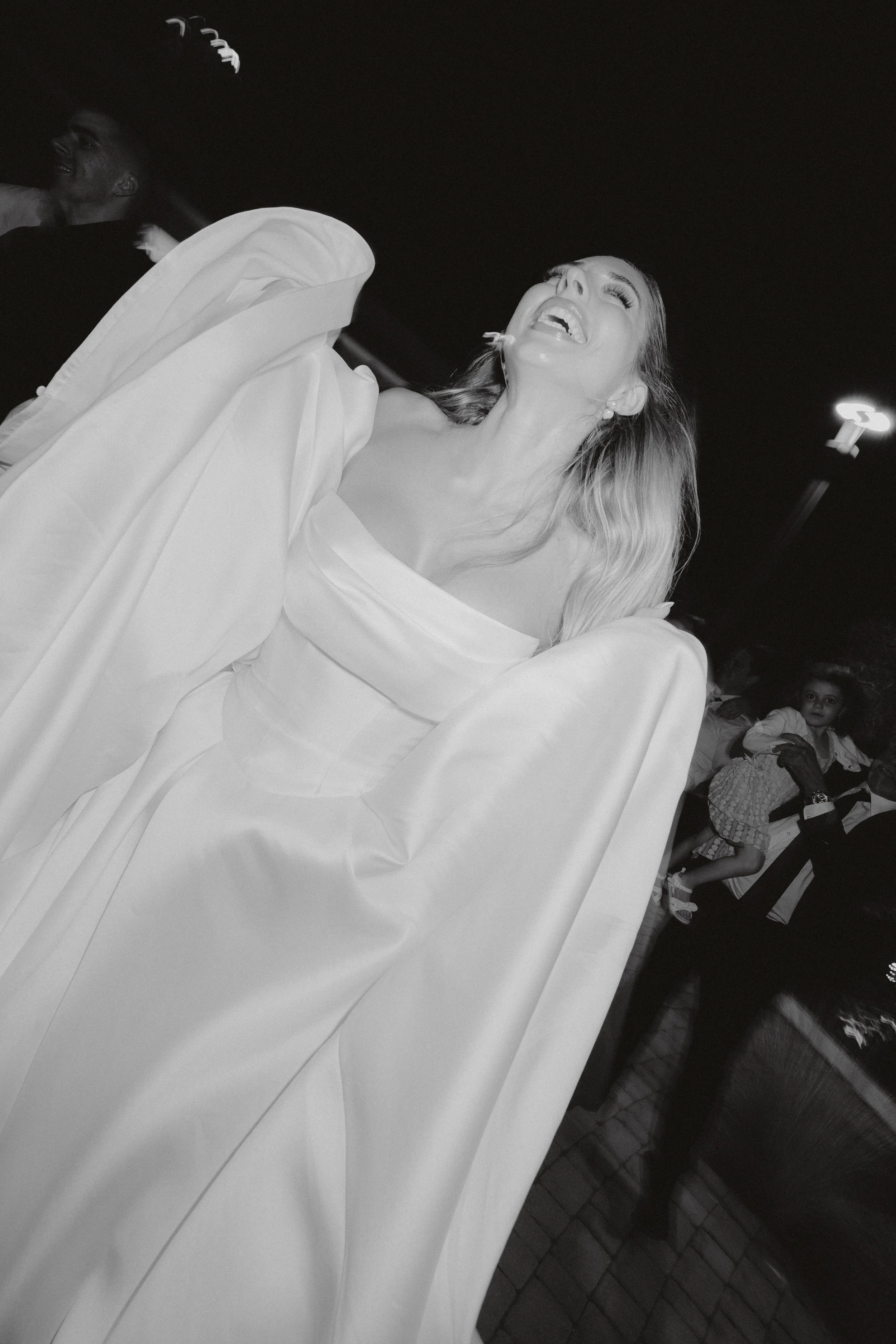 A woman with long hair laughing, wearing a white off-shoulder dress and a draped white coat, at a social gathering or party in a dimly lit room with other people in the background.