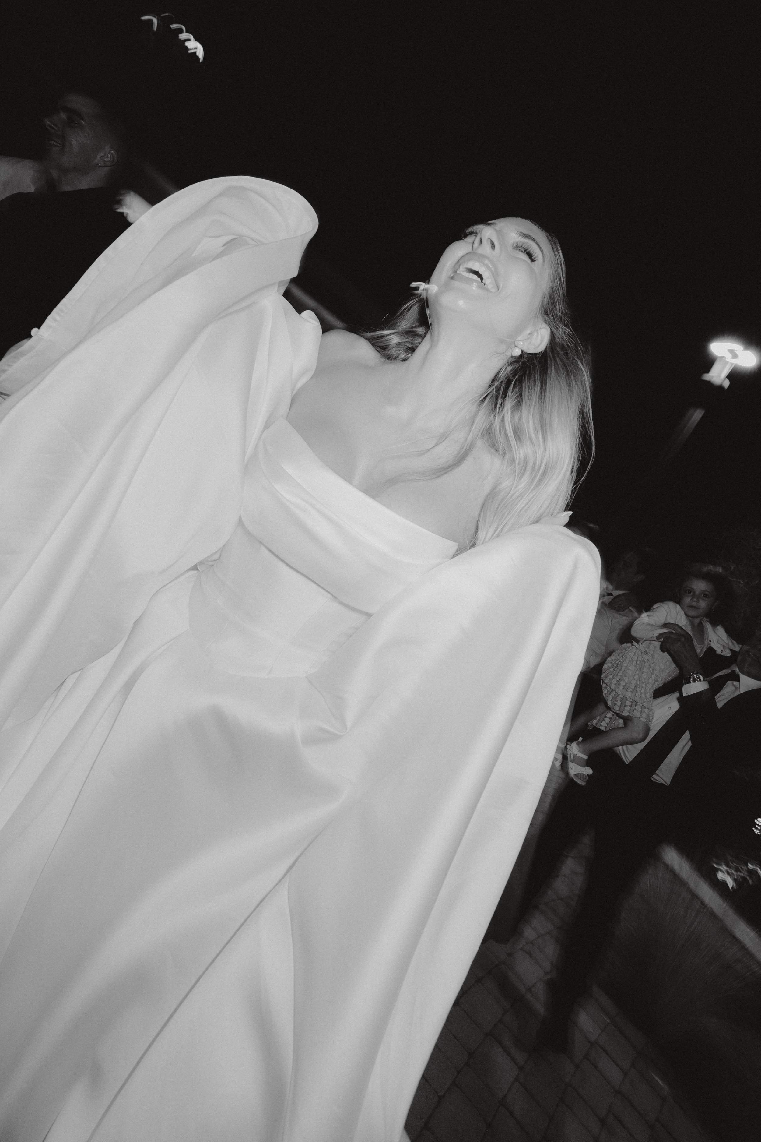 A woman in a white dress smiling and laughing, with others and children in the background.