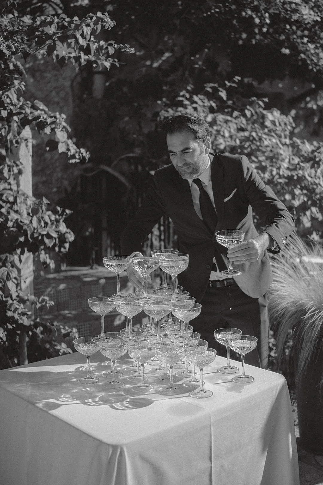 A man in a suit pours champagne into a tower of coupe glasses outdoors, with trees in the background.