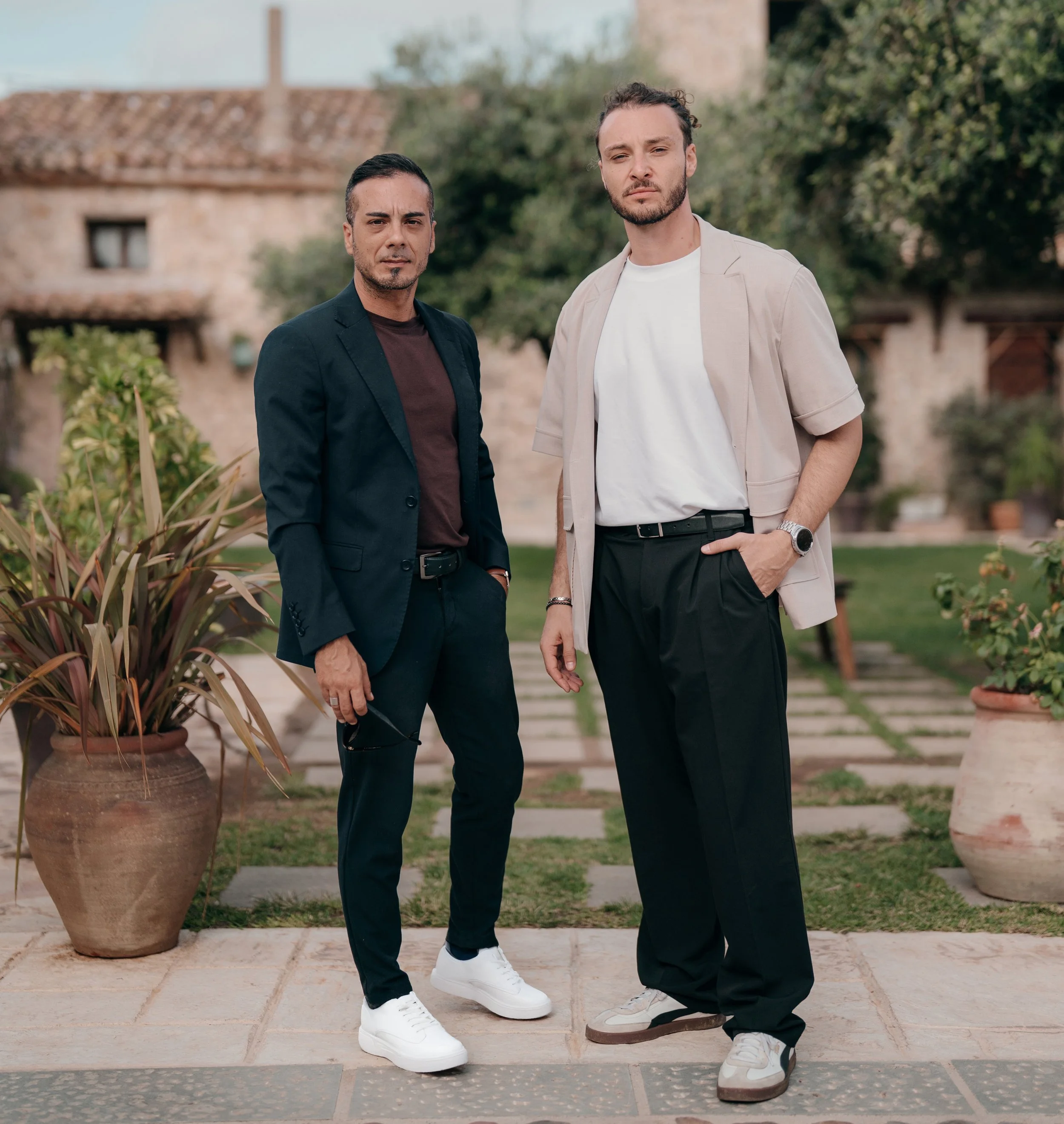 Two photographers standing outdoors on a stone pathway with potted plants and greenery, one in a black suit and the other in a beige shirt and black pants.