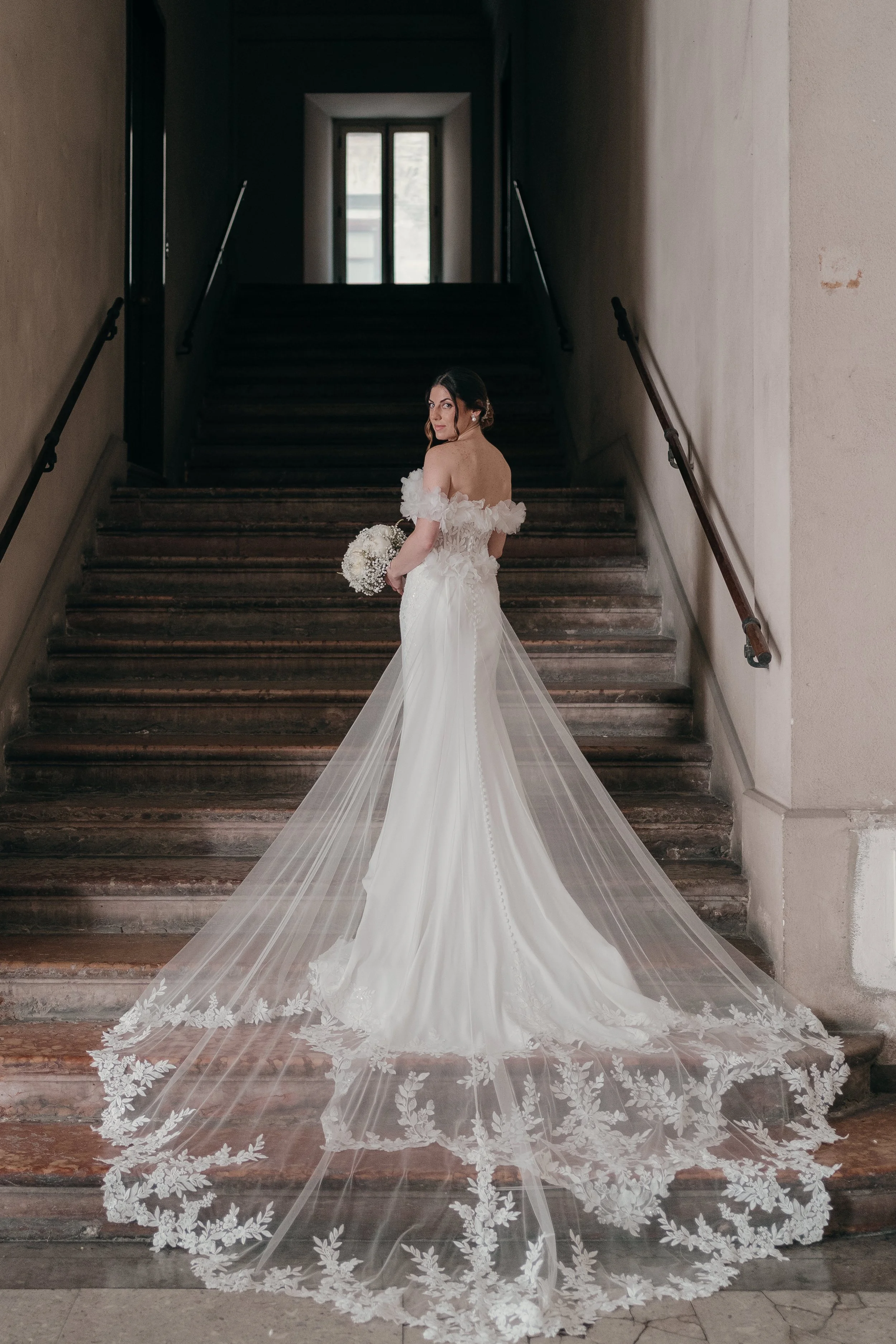 Bride in a strapless wedding dress with a long, embroidered train, standing on a staircase in a vintage building, holding a bouquet of white flowers, looking over her shoulder.