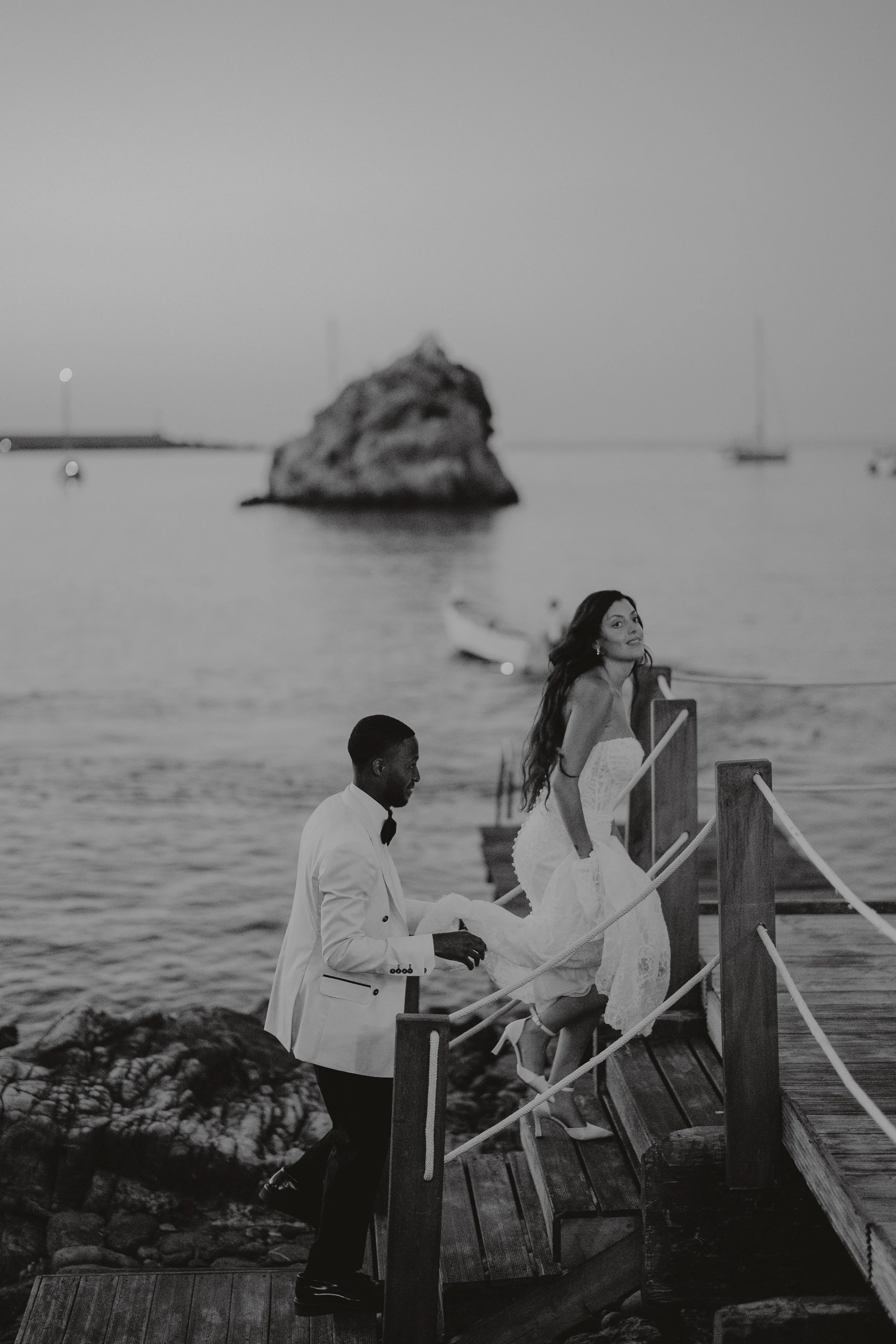 A couple dressed in wedding attire, a bride in a white dress and a groom in a tuxedo, on a wooden dock by the water with sailboats and a large rock in the background.
