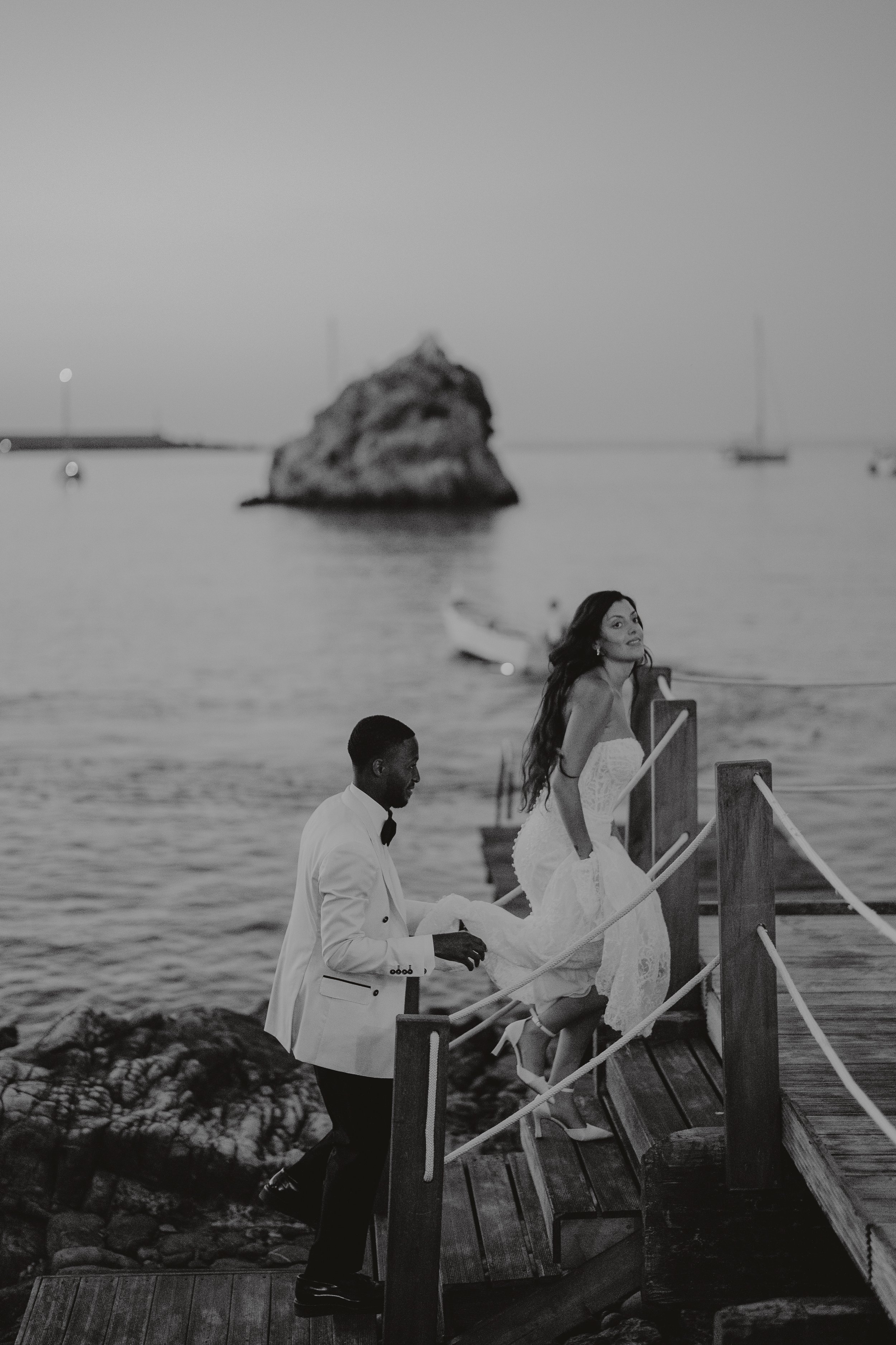 A black and white photograph of a woman in a wedding dress ascending a small wooden dock with a man in a white tuxedo holding her train, against a backdrop of a calm harbor with boats and a large rock formation in the distance.