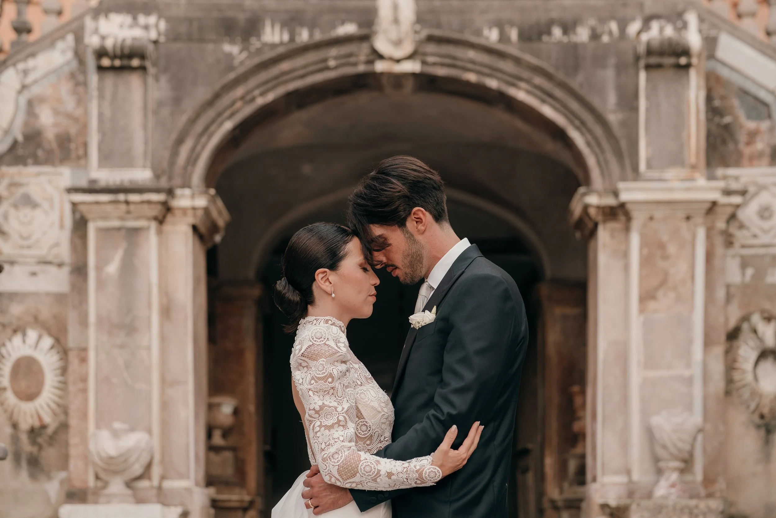 A bride and groom standing close together, touching foreheads in a romantic pose in front of an ornate stone architectural background.
