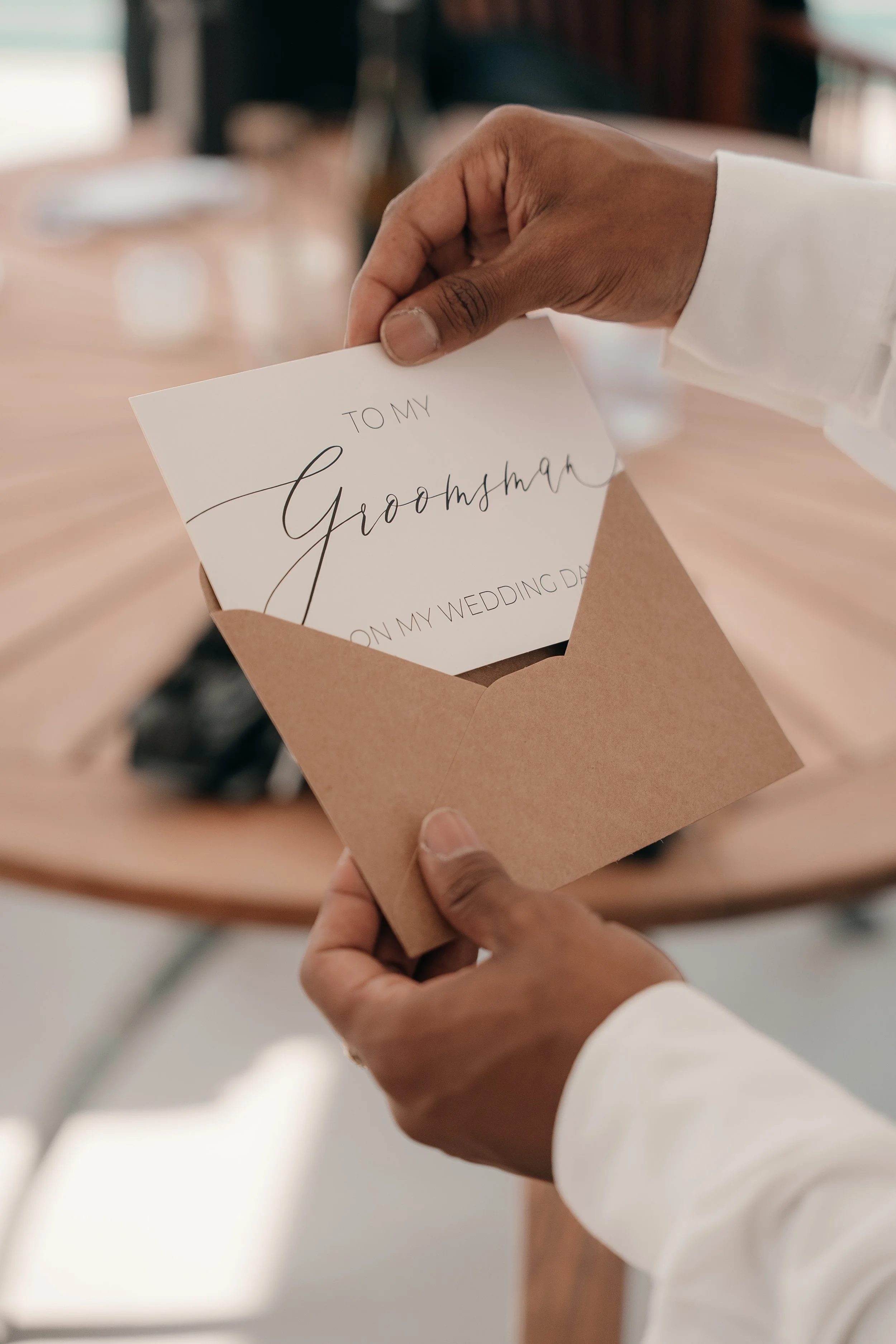 A person holding an envelope containing a card that reads "To my Groomsm" with the partial words "On my wedding dat" visible. The person's hands are visible, wearing a white long-sleeve shirt, and the background shows a wooden table.