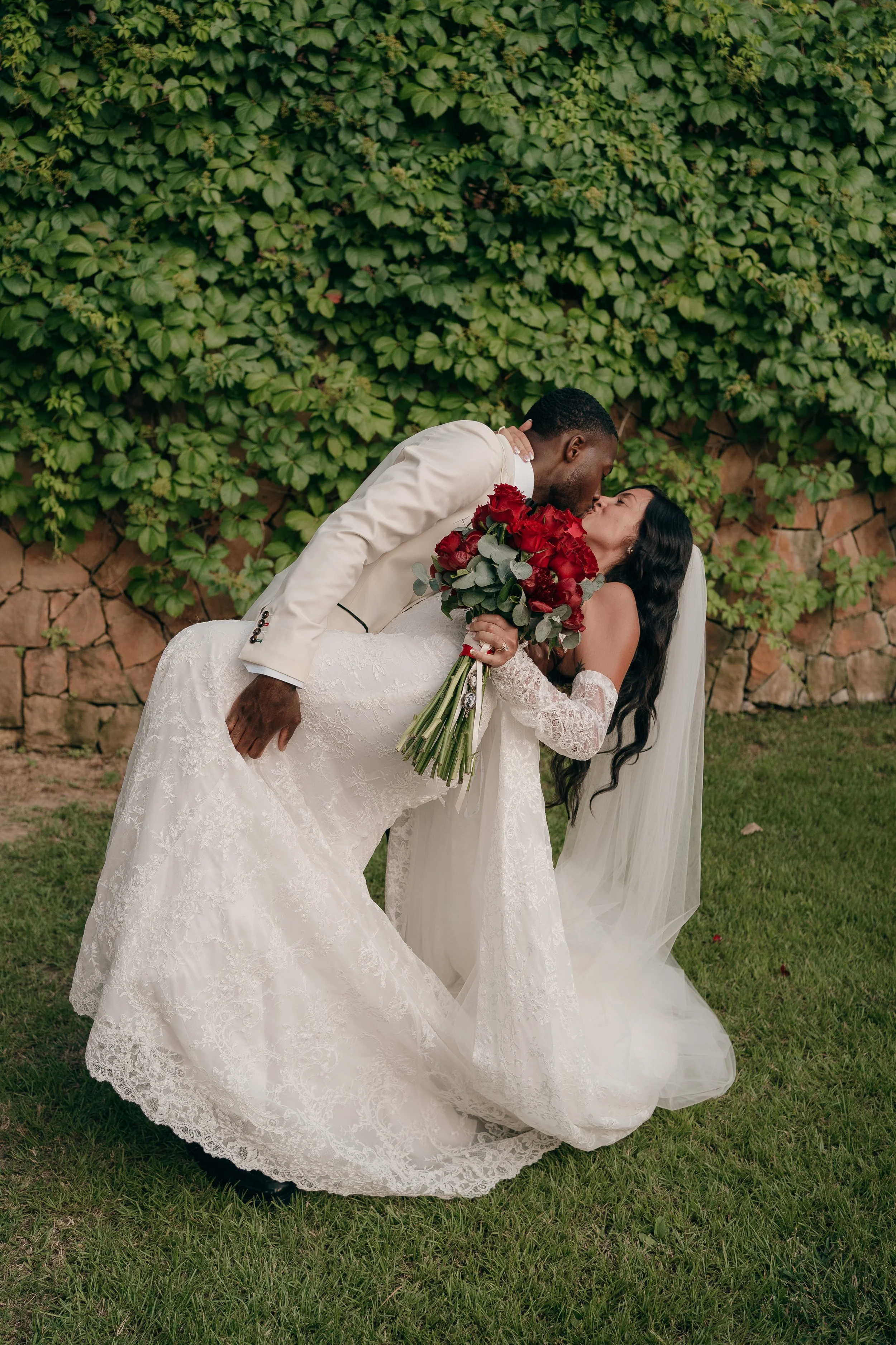 A newlywed couple sharing a romantic kiss outdoors, the groom dipping the bride while she holds a bouquet of red roses, with green foliage and stone wall in the background.