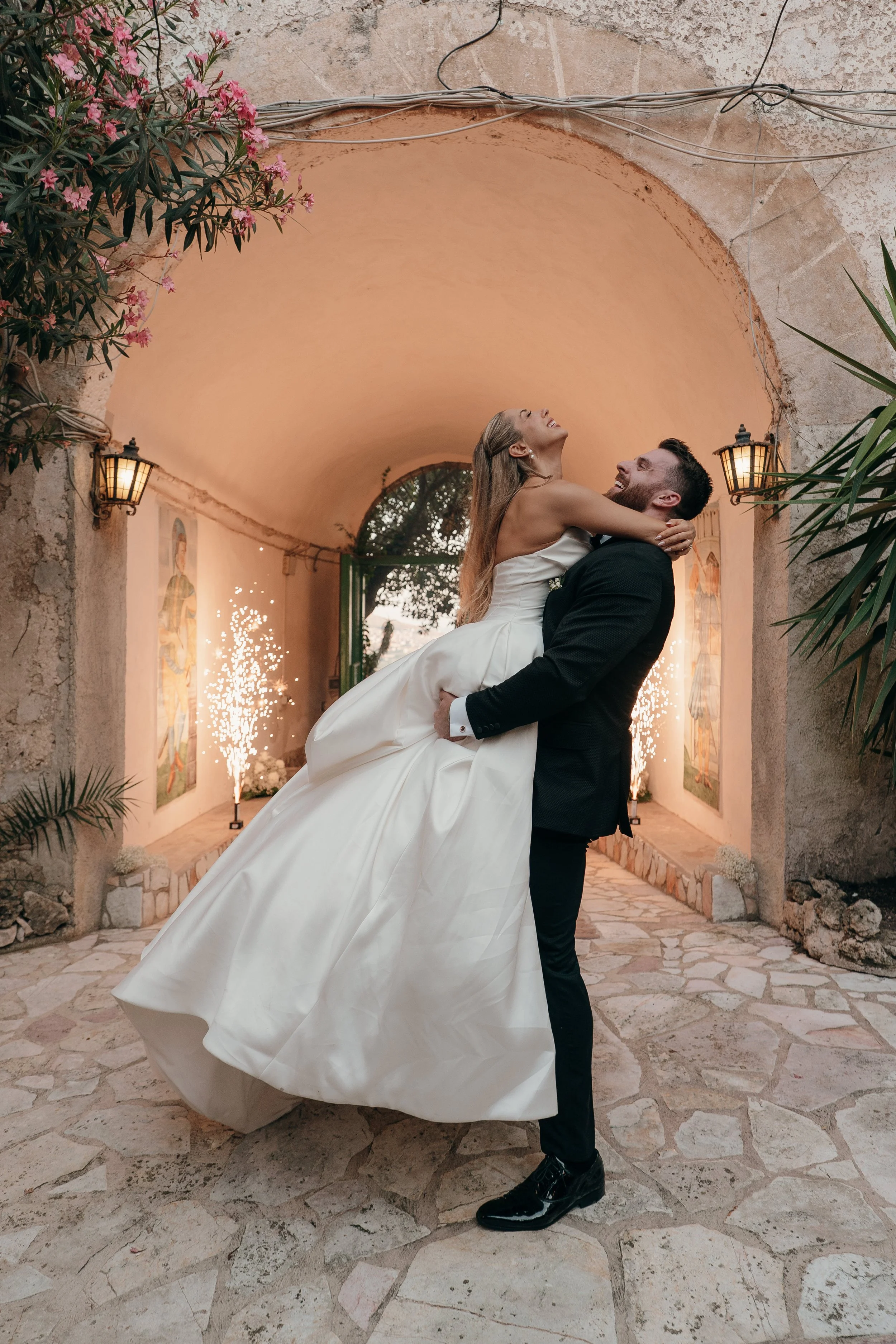 A bride being lifted by a groom inside a decorated venue with lights and artwork on the walls.