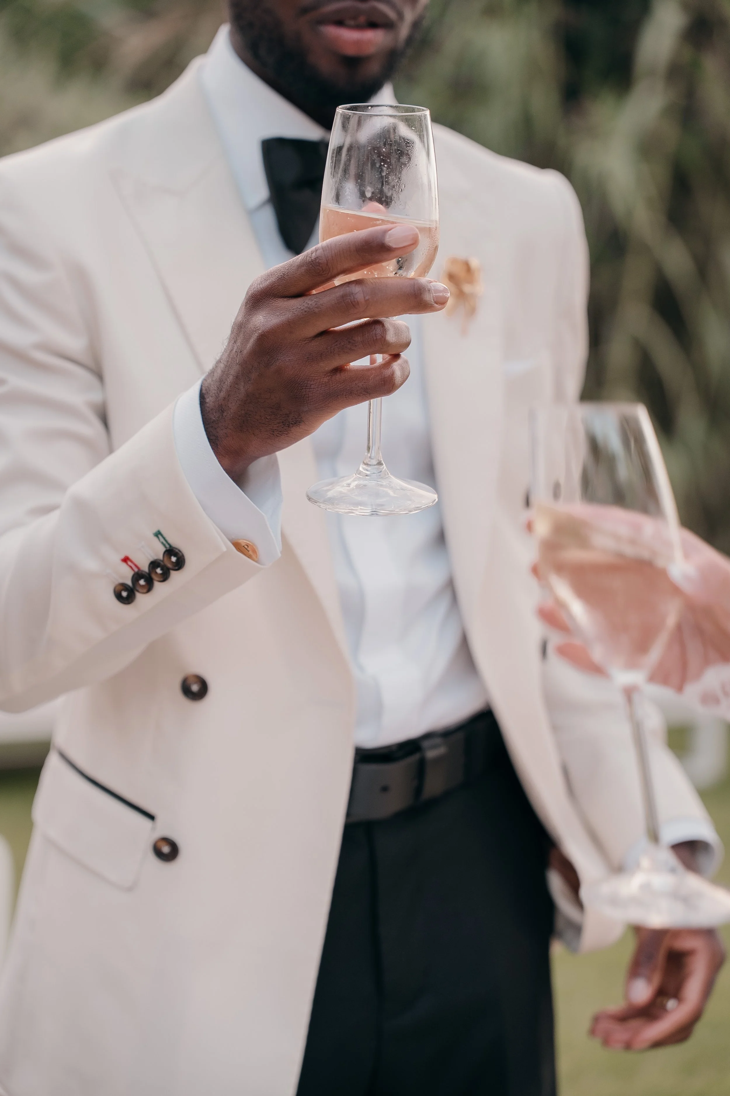 Man in a white tuxedo holding a glass of champagne at an outdoor event.
