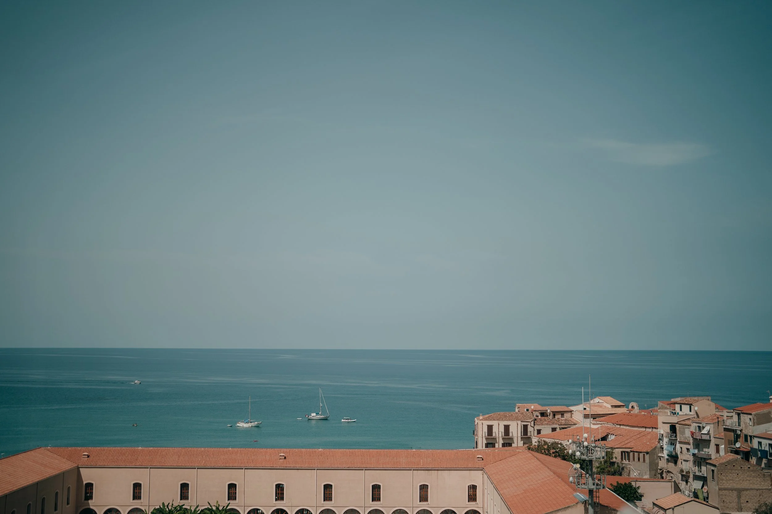 A seaside town with orange-roofed buildings overlooking the ocean. Several sailboats are anchored in the water. The sky is clear with a few wispy clouds.