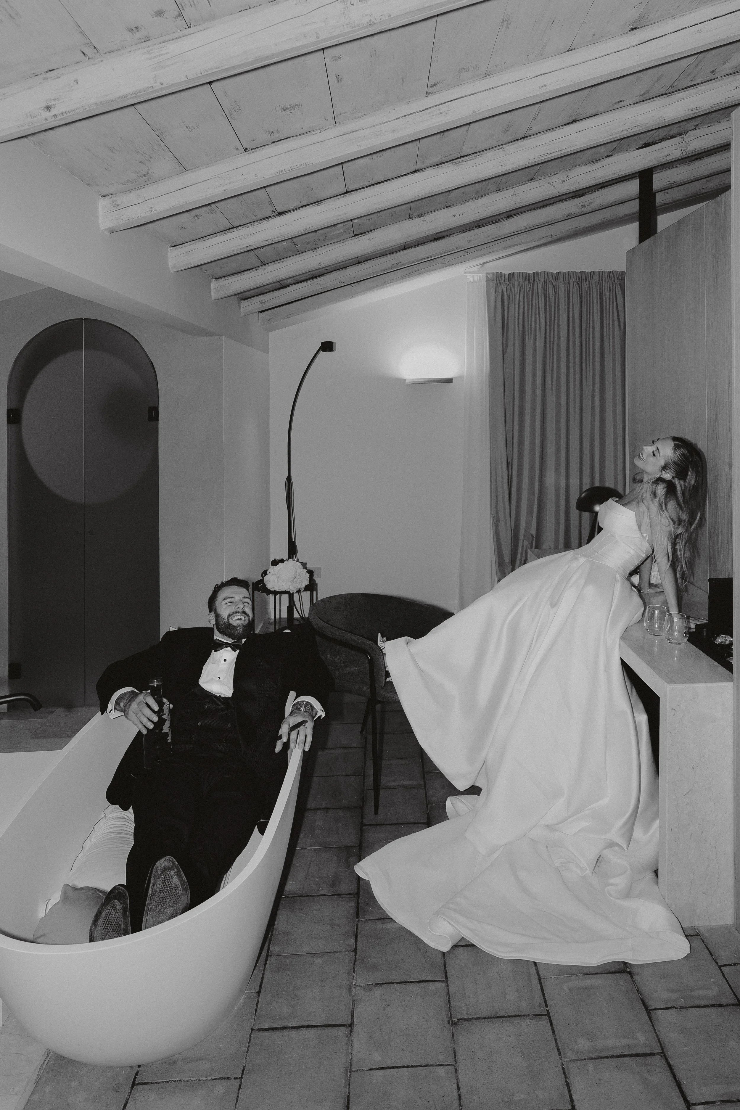 A black and white photo of a man in a tuxedo relaxing in a modern bathtub, with a woman in a wedding dress sitting on a counter behind him, all in a stylish room with wooden ceiling beams and simple decor.