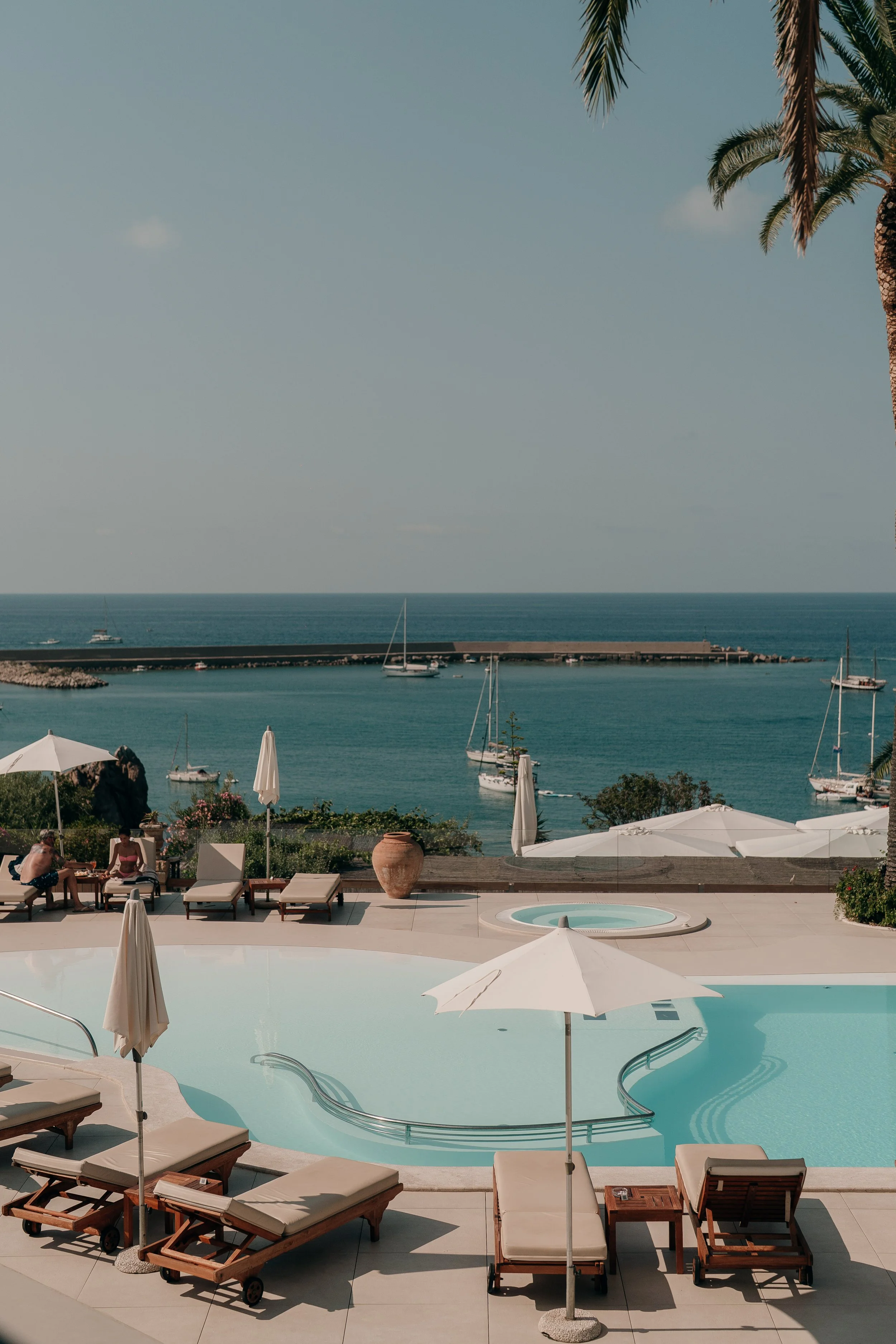 Poolside area with lounge chairs, umbrellas, and people, overlooking the ocean with sailboats and a breakwater