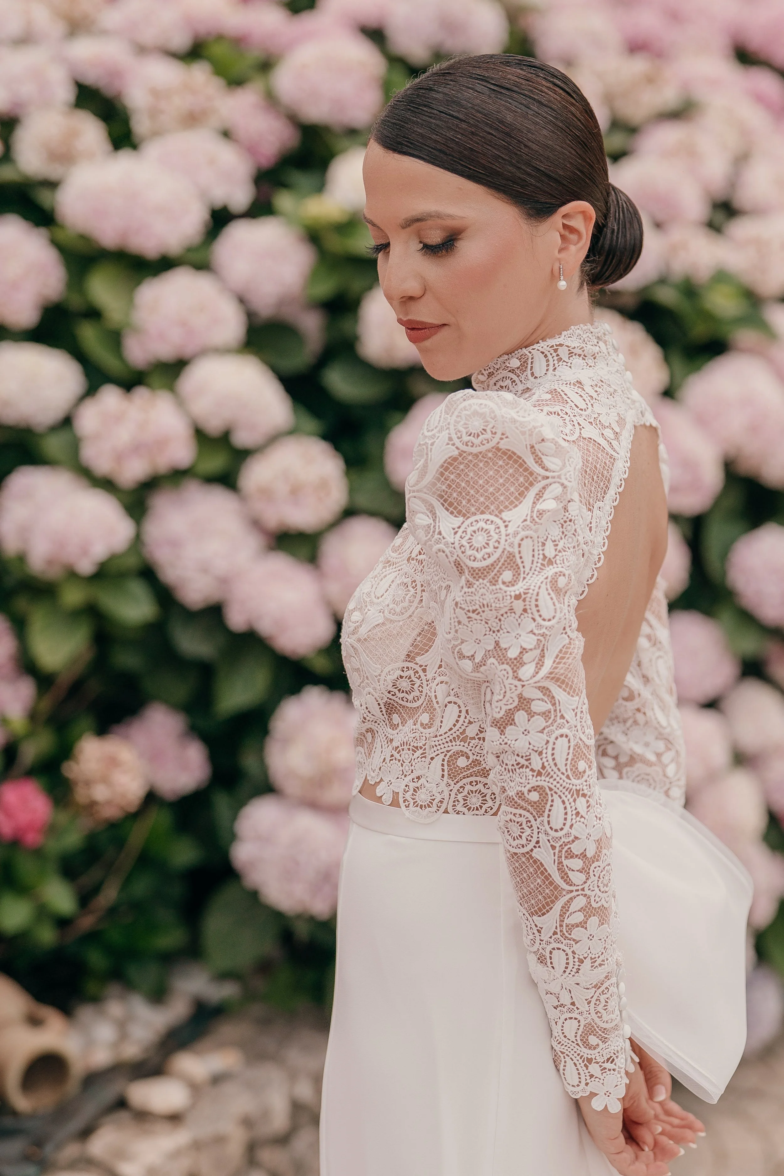 A woman in a white lace top and white skirt, holding a white clutch, stands against a background of pink hydrangea flowers.
