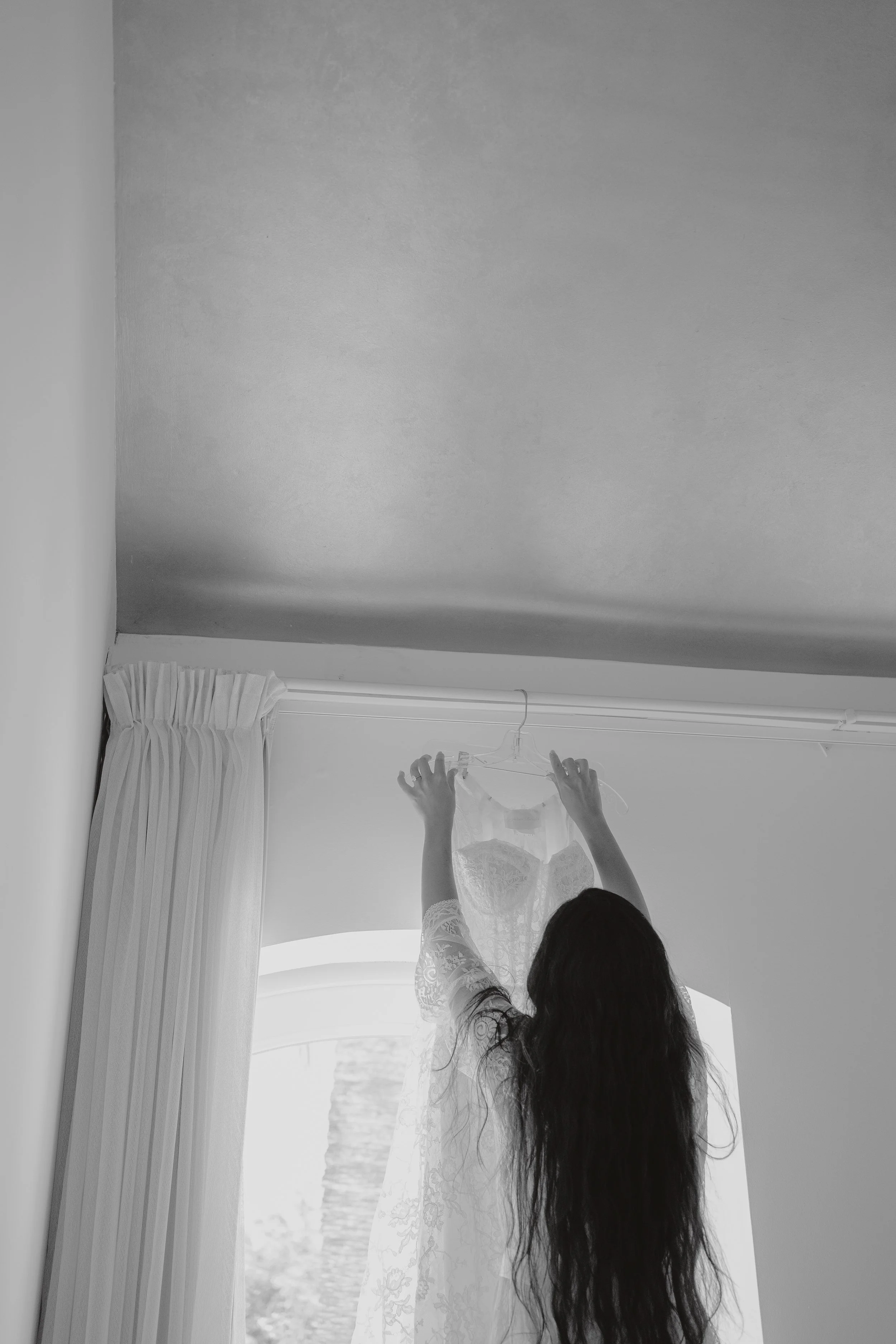 A woman with long dark hair, viewed from behind, holding a lace wedding dress up to hang it. She is inside a room near a window with curtains.