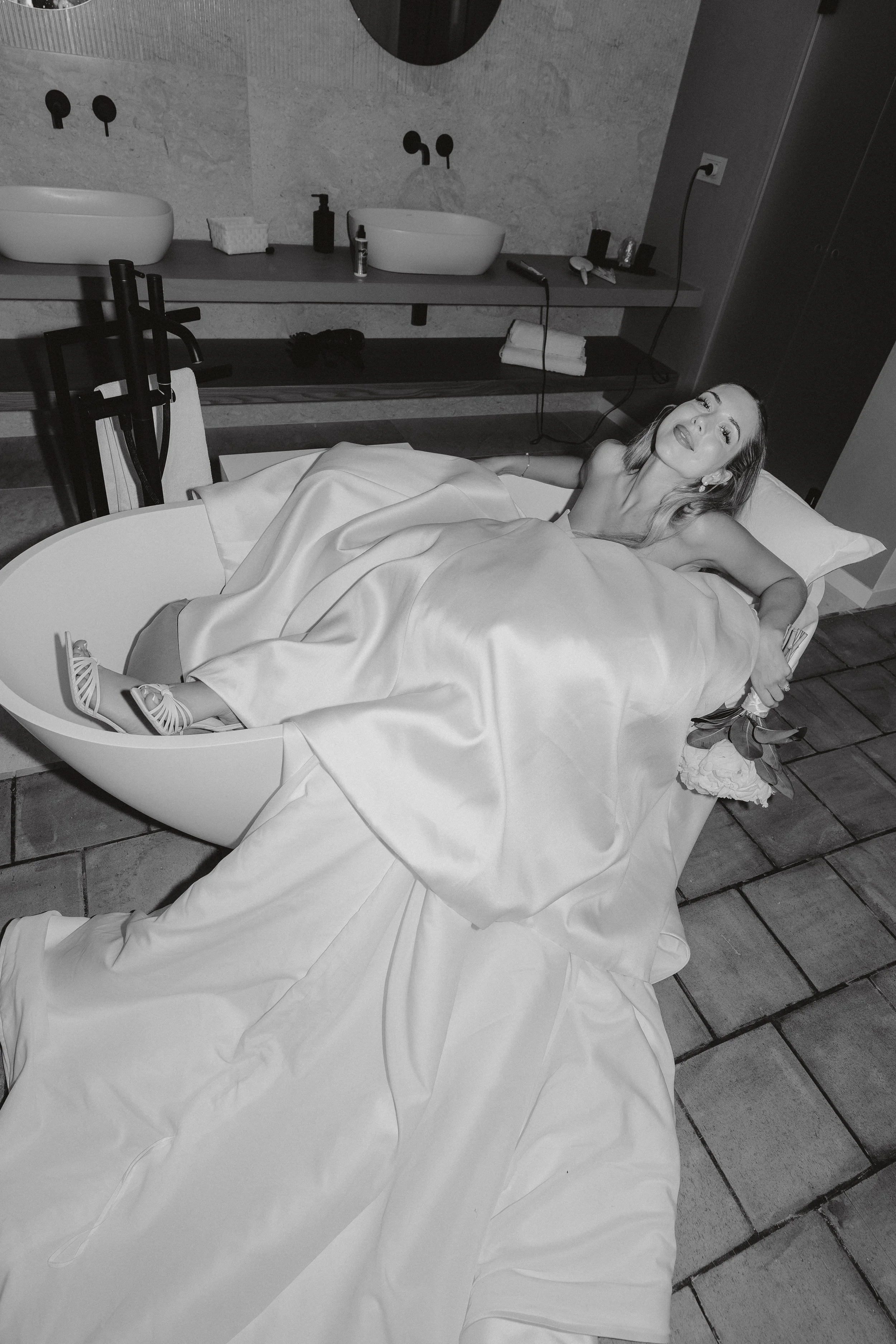 A woman in a wedding dress lying on a lounge chair in a modern bathroom.