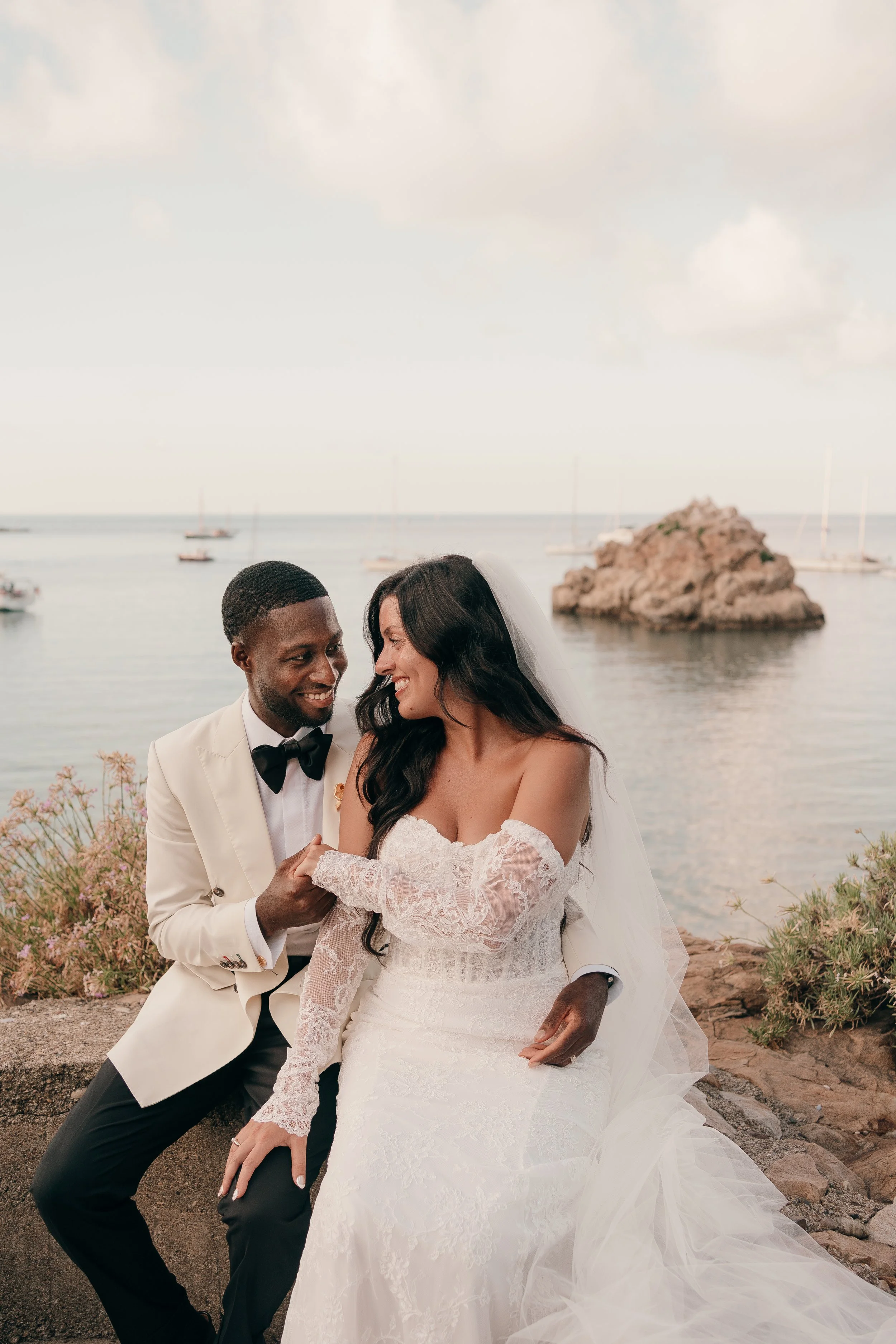 A bride and groom sitting together by the water at a seaside wedding, smiling and holding hands. The bride is in a white lace wedding dress, and the groom is in a white tuxedo jacket with black pants and a bow tie. There are boats and rocks in the ba