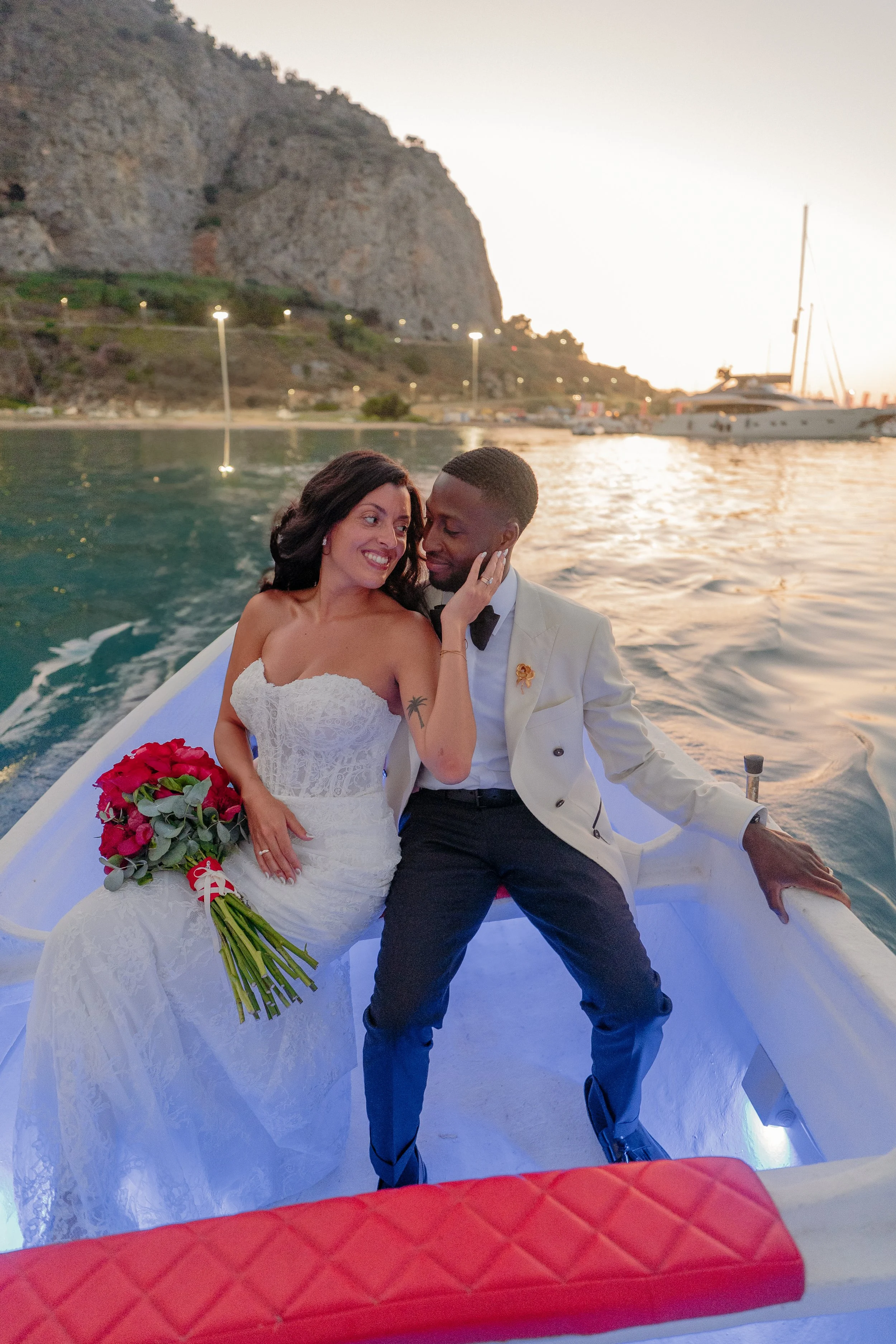 A newlywed couple in wedding attire, sitting in a boat on the water at sunset, with a rocky hillside and sailboats in the background.