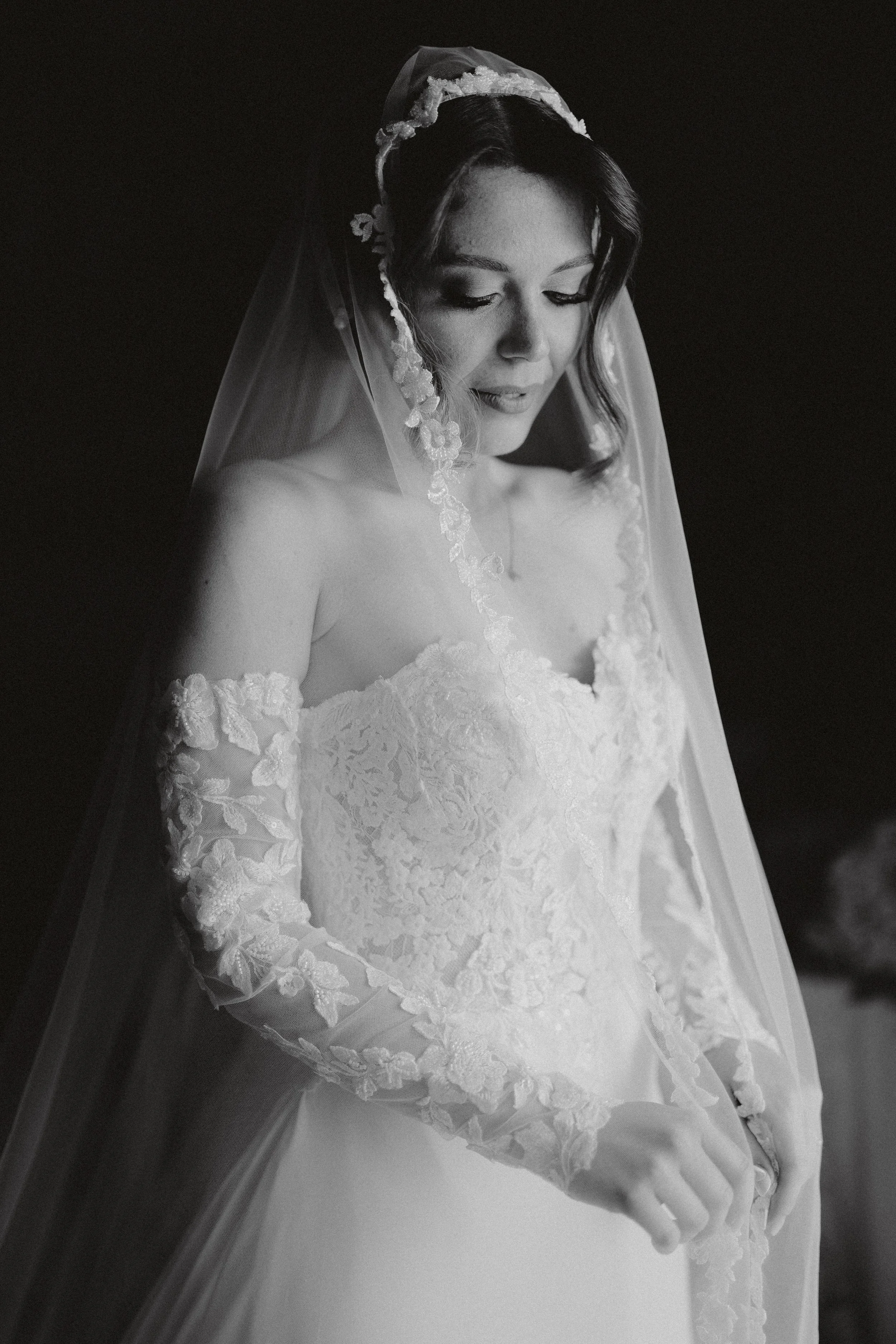 Black and white photograph of a bride in a lace wedding gown with long lace sleeves, wearing a veil with floral accents, looking down with a peaceful expression.
